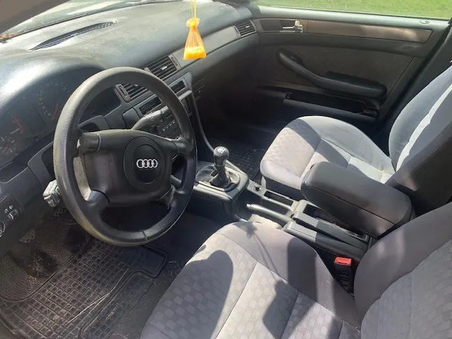 Wnętrze samochodu marki Audi z perspektywy kierowcy. Widoczna jest czarna kierownica z logo Audi w centrum, deska rozdzielcza, dźwignia zmiany biegów oraz dwa szare, materiałowe fotele. Na lusterku wstecznym wisi żółty odświeżacz powietrza. Podłoga wyłożona jest czarnymi gumowymi dywanikami.