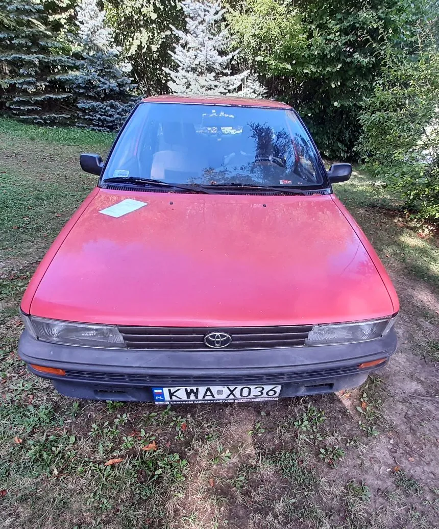 Widok z przodu na czerwony samochód marki Toyota zaparkowany na trawiastym podłożu. Pojazd posiada ciemnoszarą przednią maskę i zderzak. Na zderzaku znajduje się polska tablica rejestracyjna o numerze KWA X036. W tle widoczne są zielone drzewa i krzewy.
