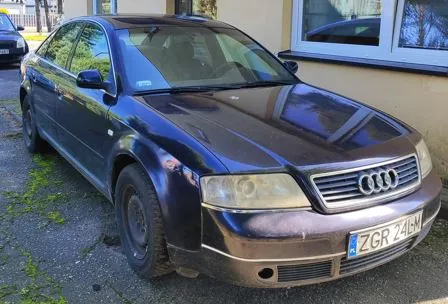 Ciemnofioletowy samochód marki Audi zaparkowany na podjeździe przed jasnym budynkiem. Widoczny jest przód i bok auta, grill z logo Audi oraz tablica rejestracyjna o numerze ZGR 24LM.