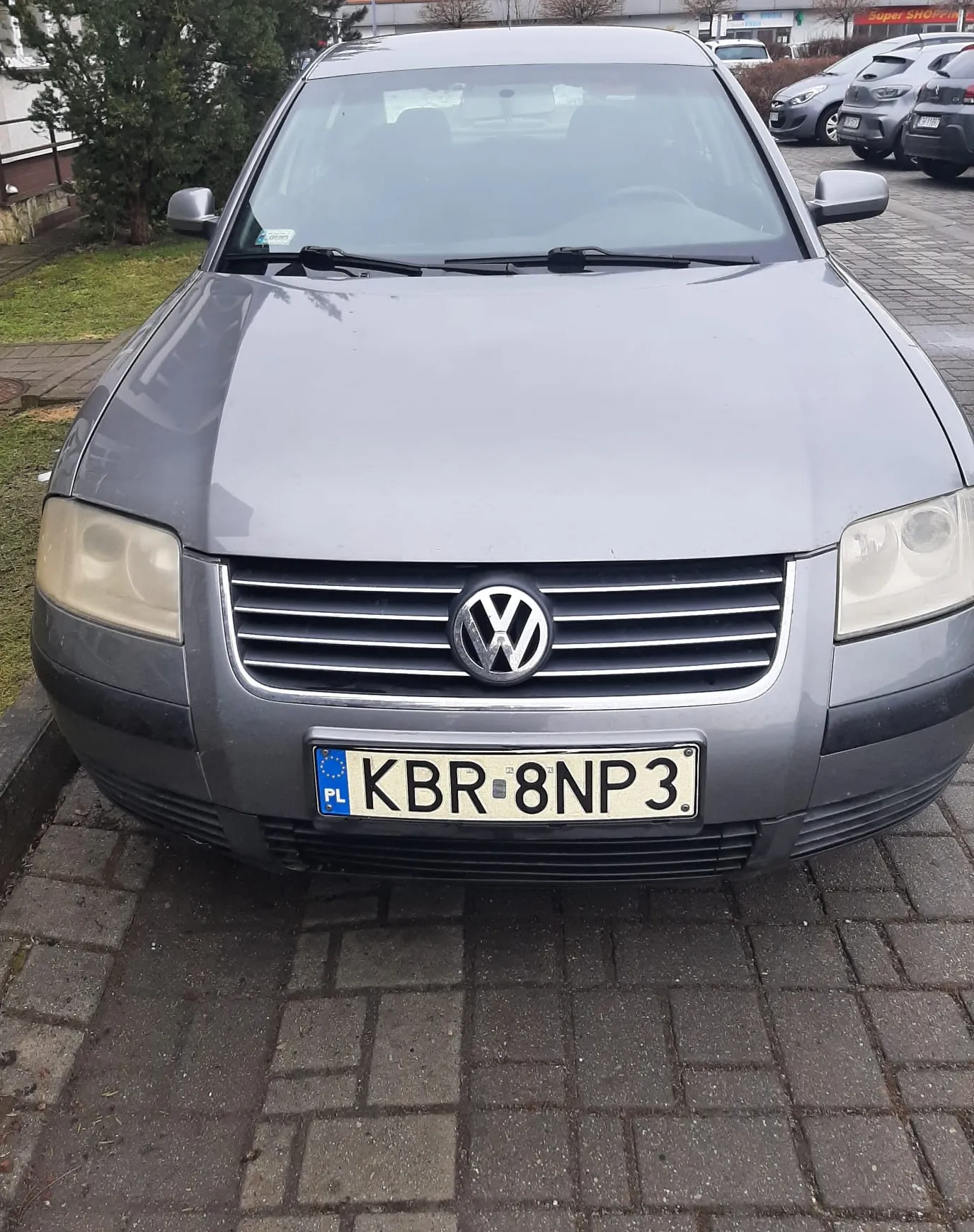 Przedni widok szarego samochodu marki Volkswagen zaparkowanego na brukowanej powierzchni. Na masce widoczny jest logo VW, a poniżej tablica rejestracyjna z białym tłem, niebieskim pasem po lewej stronie z napisem PL oraz czarnym tekstem KBR 8NP3. W tle znajdują się inne zaparkowane samochody, zieleń, drzewa oraz budynki z widocznym napisem Super SHOP. Samochód stoi na chodniku z kostki brukowej, po lewej stronie widoczna trawa i krzewy.
