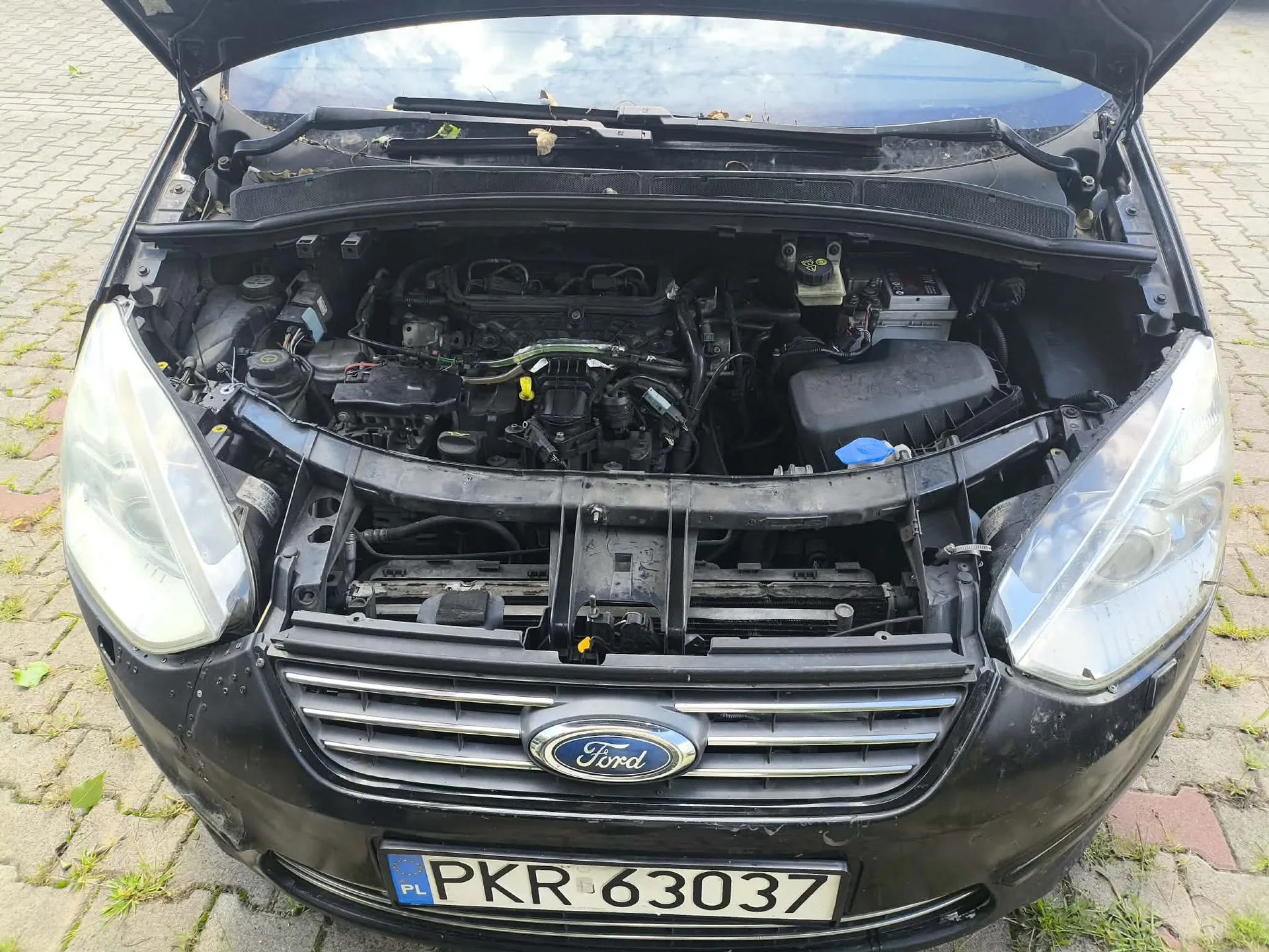 Widok z przodu na czarny samochód marki Ford z otwartą maską, odsłaniającą silnik i komponenty mechaniczne. Na przednim zderzaku znajduje się polska tablica rejestracyjna o numerze PKR 63037.