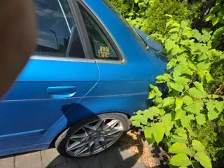 Zbliżenie na tylną część niebieskiego samochodu zaparkowanego obok zielonego krzewu. Na tylnej szybie widoczna jest naklejka z tekstem 'JDM TURBO'. Widoczna jest srebrna felga z wieloramiennym wzorem.