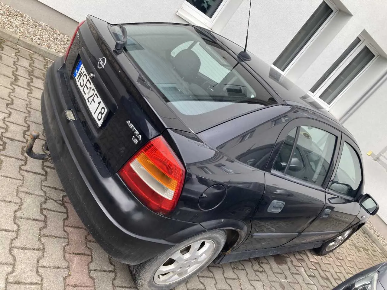 Czarny samochód Opel Astra z tablicą rejestracyjną WSE 2F18 zaparkowany na brukowanej powierzchni przed białym budynkiem z oknami. Widoczne są tylna część pojazdu, czerwone światło tylnego, a na bagażniku napis 'Astra' oraz logo marki.