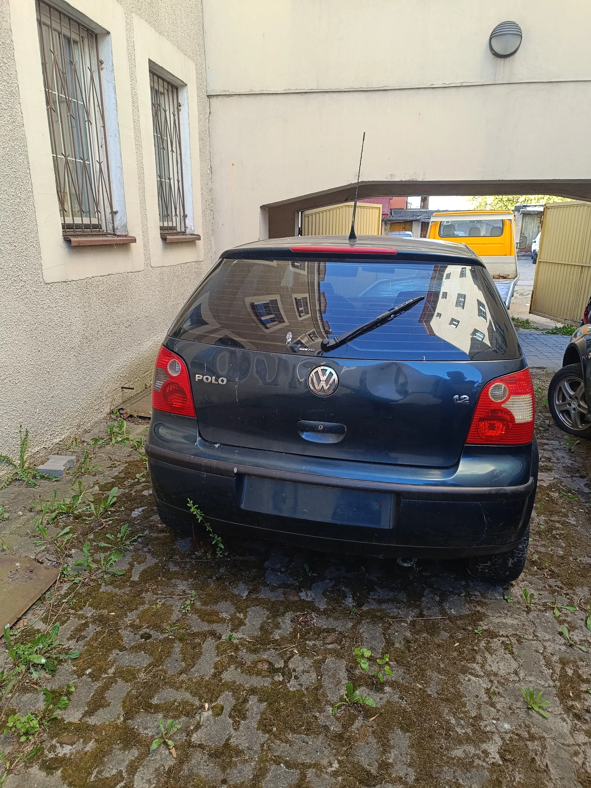 Czarny samochód Volkswagen Polo z tyłu, z widocznym logo VW, napisem "POLO" po lewej stronie bagażnika i oznaczeniem "1.2" po prawej. Brak tablicy rejestracyjnej. Samochód zaparkowany na betonowej powierzchni z mchem i roślinami. W tle budynek z jasną elewacją, dwoma oknami z metalowymi kratami po lewej stronie oraz przejazdem prowadzącym do innego obszaru, gdzie widoczny jest żółty samochód. Po prawej stronie fragment innego pojazdu.