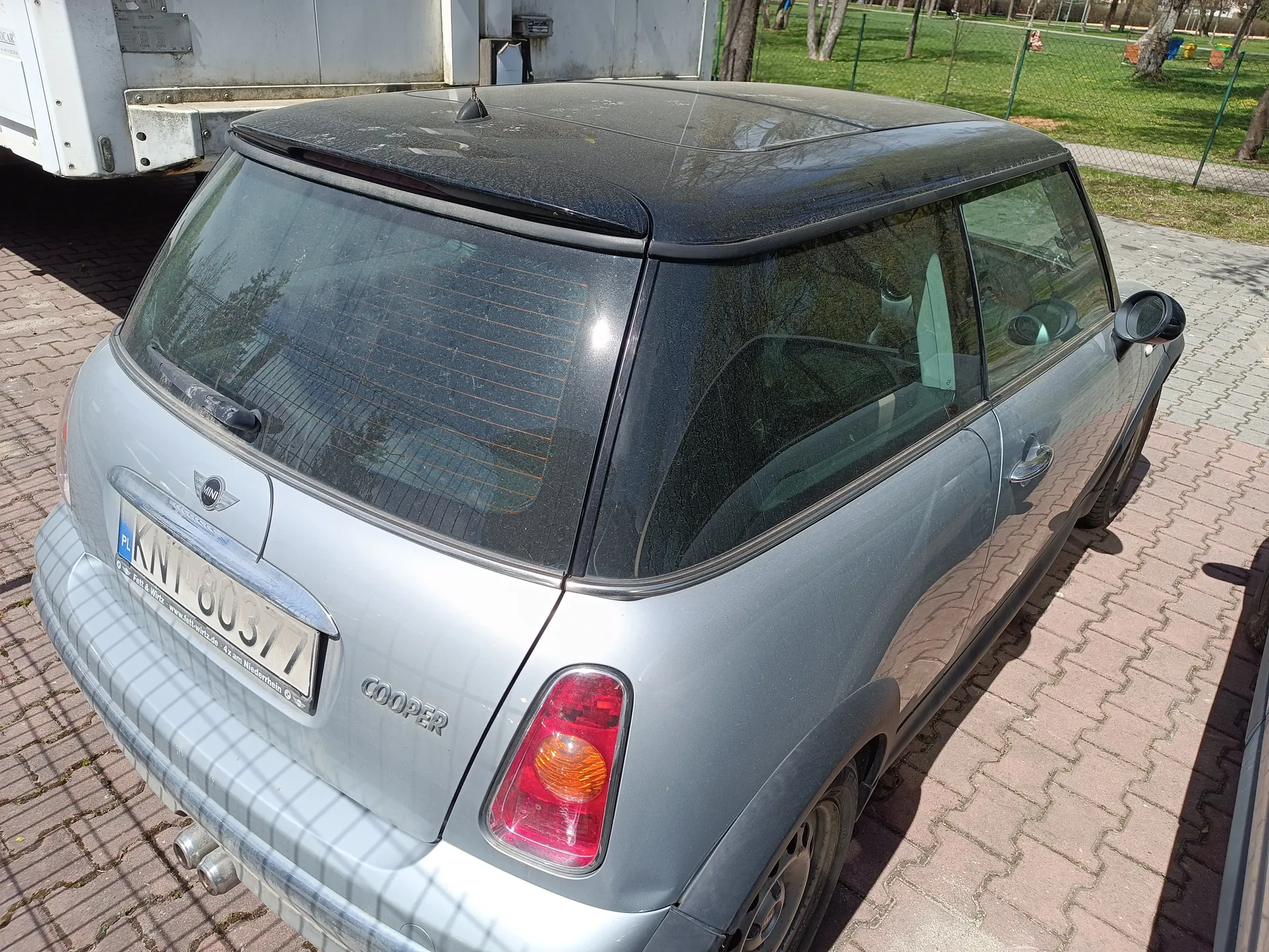 Srebrny samochód Mini Cooper z widocznym tyłem, tablicą rejestracyjną PL KNT 80377 i napisem COOPER na bagażniku. Samochód zaparkowany na brukowanej powierzchni, w tle biały przyczepa, trawnik, ogrodzenie i drzewa. Słoneczny dzień, widoczne cienie na ziemi.