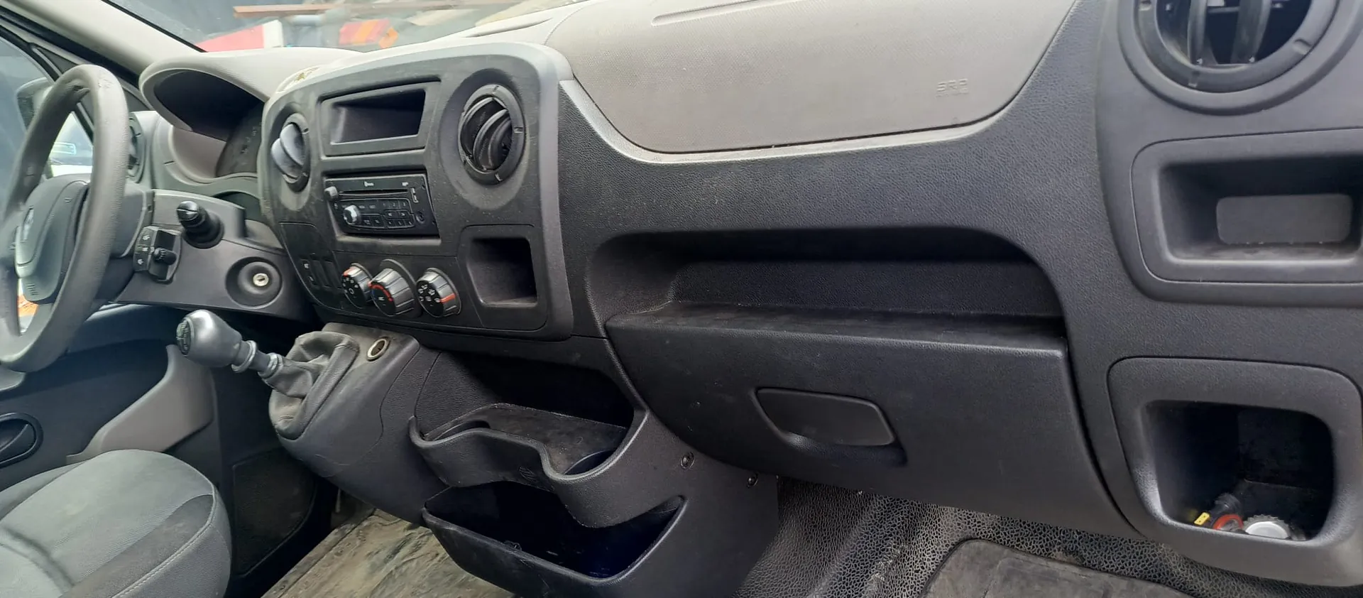 Wnętrze samochodu z widokiem na deskę rozdzielczą wykonaną z ciemnoszarego plastiku. Widoczna jest kierownica po lewej stronie, konsola środkowa z radiem, pokrętłami klimatyzacji oraz nawiewami powietrza. Poniżej znajduje się dźwignia zmiany biegów oraz schowki. Po prawej stronie widoczny jest schowek dla pasażera i dodatkowy uchwyt.