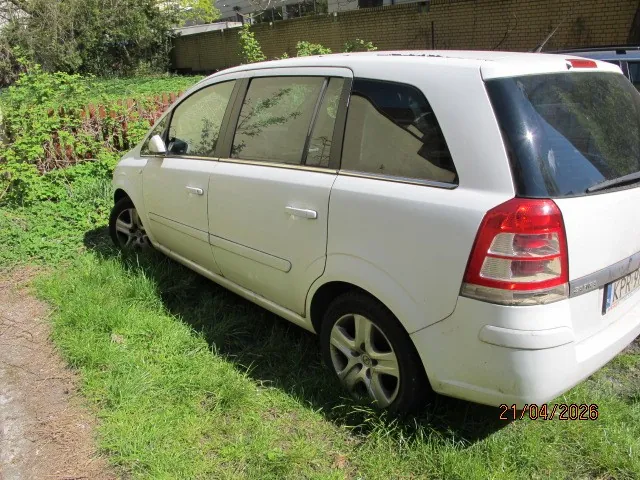 Biały samochód typu kombi zaparkowany na trawie. Widoczna tylna część pojazdu z czerwonymi światłami i częściowo czytelną tablicą rejestracyjną z napisem KEX. W tle zielona trawa, krzewy, ceglany mur oraz ogrodzenie. W prawym dolnym rogu obrazu znajduje się data 21/04/2023.