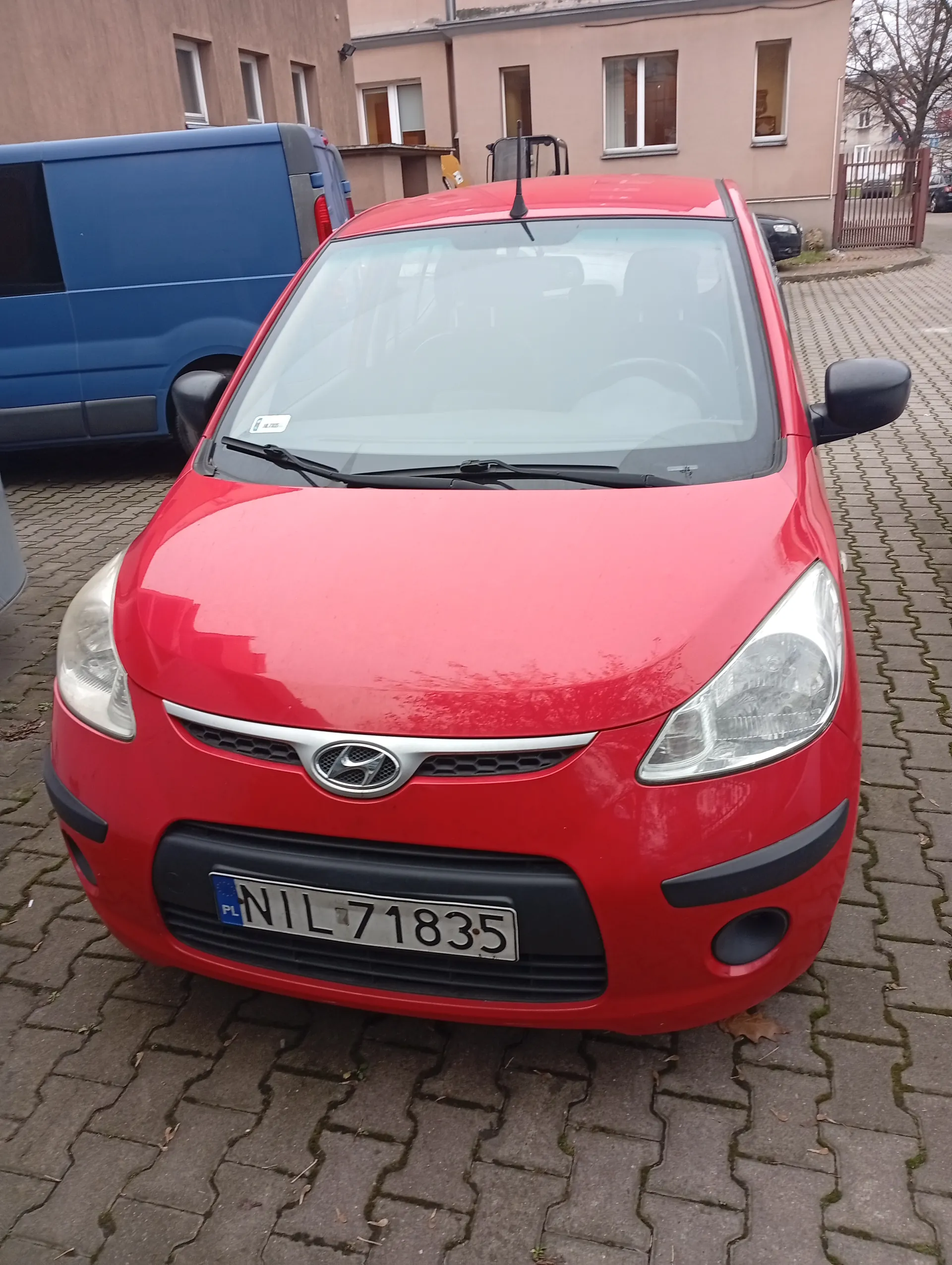 Czerwony samochód marki Hyundai z tablicą rejestracyjną PL NIL 71835 zaparkowany na brukowanej powierzchni. W tle widoczny niebieski furgon oraz budynki mieszkalne z oknami i bramą. Samochód ma czarne zderzaki, reflektory i logo marki na masce. Na przedniej szybie znajduje się biała naklejka.