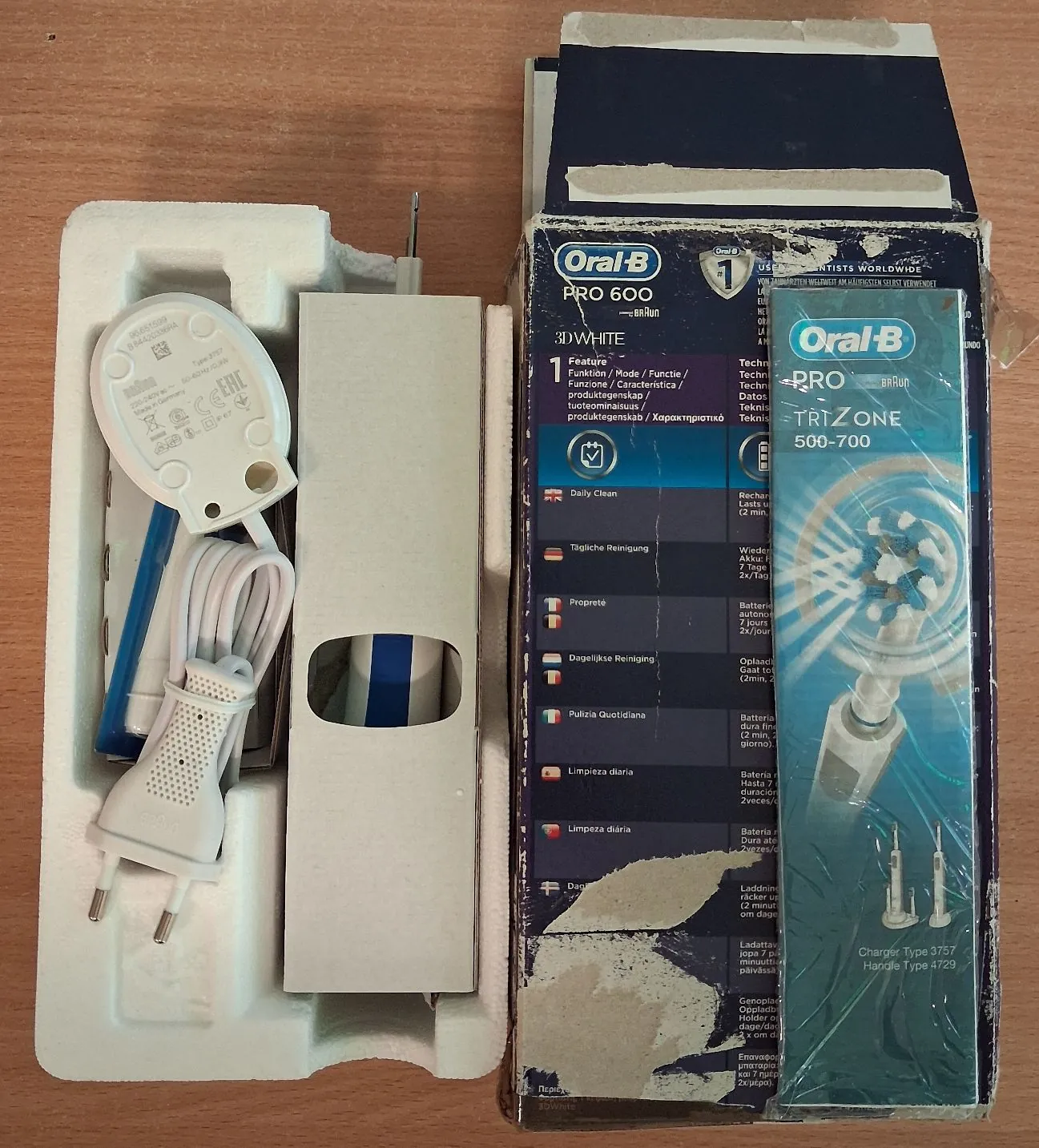Zdjęcie zestawu szczoteczki elektrycznej Oral-B PRO 600. Po lewej stronie znajduje się biała styropianowa tacka z białym zasilaczem i kablem. Po prawej stronie stoi niebiesko-białe opakowanie kartonowe z logo Oral-B, nazwą modelu PRO 600 oraz napisem TRIZONE 500-700. Na pudełku widoczne są opisy funkcji w kilku językach oraz ilustracja końcówki szczoteczki.