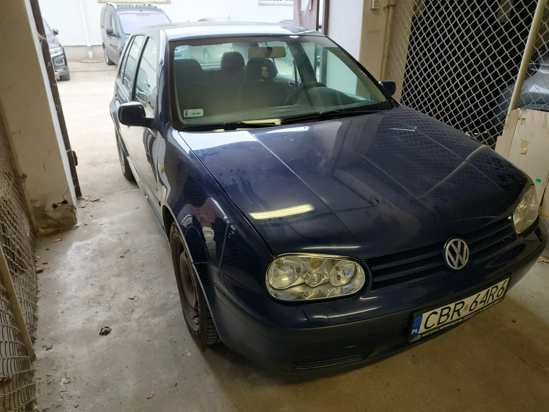 Ciemnoniebieski samochód Volkswagen Golf zaparkowany w garażu. Widoczna tablica rejestracyjna z polskim kodem 'PL' i numerem 'CBR 64R6'. W tle inne pojazdy, siatka ochronna oraz betonowa podłoga. Przez przednią szybę widać siedzenia i lusterko wsteczne.