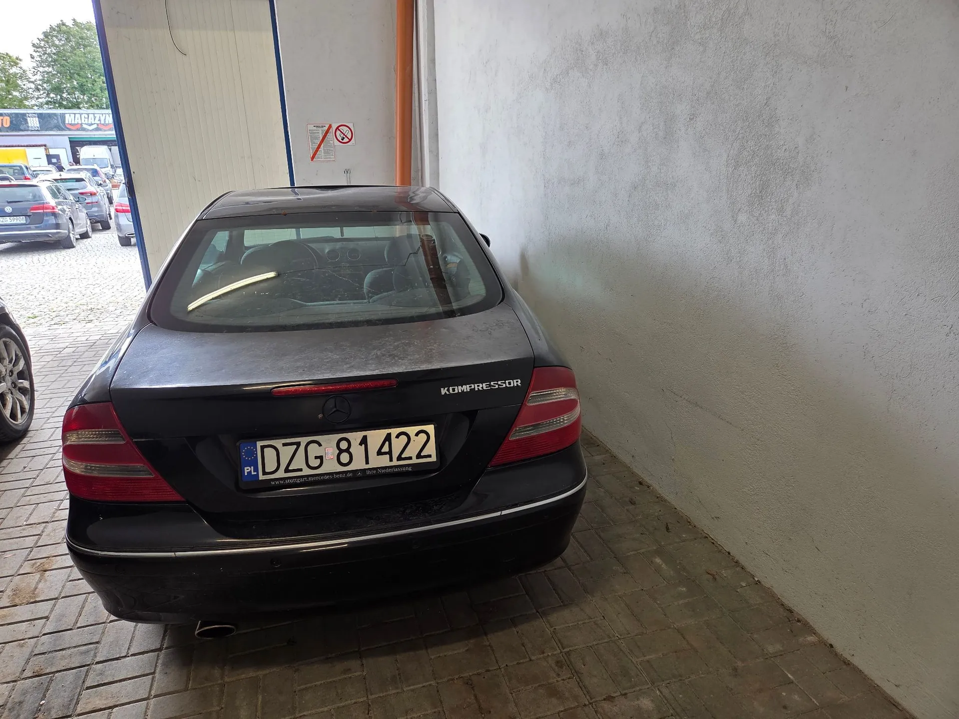 Tylny widok czarnego samochodu osobowego marki Mercedes-Benz zaparkowanego w półotwartej hali lub garażu. Samochód posiada tablicę rejestracyjną o numerze DZG 81422 oraz napis KOMPRESSOR na klapie bagażnika. Po prawej stronie znajduje się jasna, betonowa ściana, a w tle widoczny jest parking z innymi pojazdami i budynek z napisem MAGAZYN.