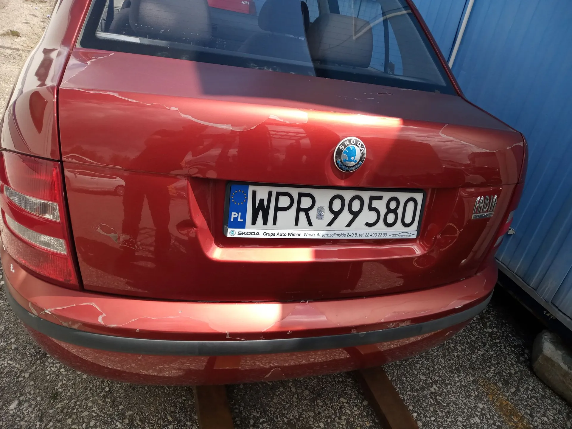 Tylny widok czerwonego samochodu marki Skoda Fabia. Pojazd posiada tablicę rejestracyjną o numerze WPR 99580. Na klapie bagażnika widoczne jest logo marki Skoda oraz napis Fabia po prawej stronie. Na ramce tablicy rejestracyjnej znajduje się tekst: 'SKODA Grupa Auto Wimar W-wa, al. Jerozolimskie 249 B, tel. 22 490 22 33'. Karoseria samochodu posiada liczne zarysowania i wgniecenia, szczególnie w okolicach tablicy rejestracyjnej i na zderzaku.