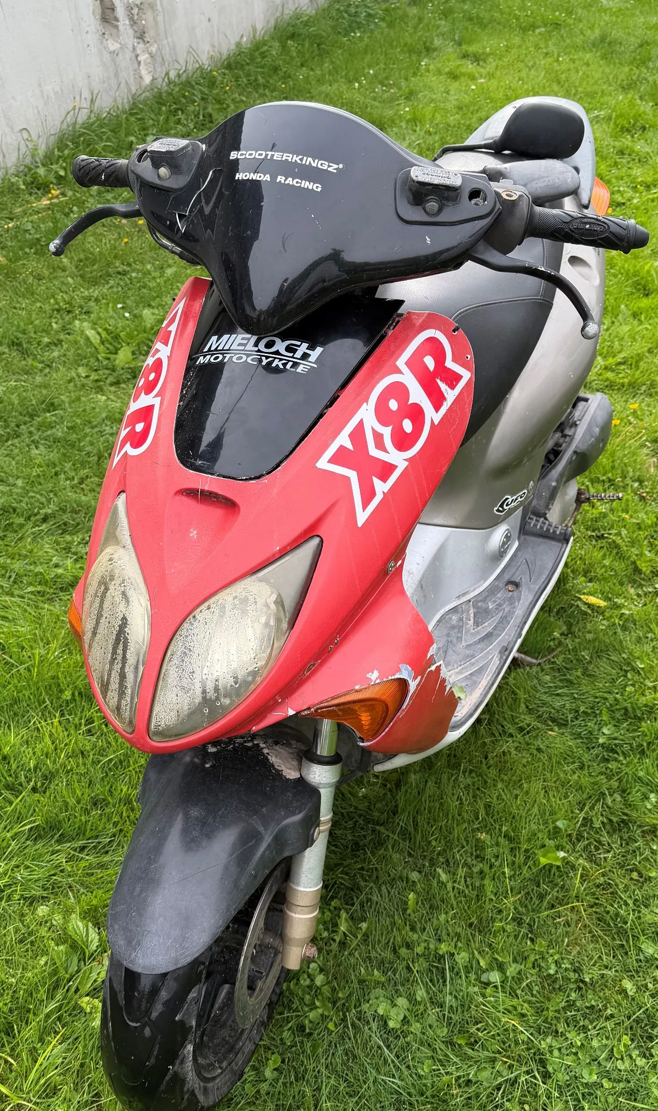 Czerwono-srebrny skuter zaparkowany na zielonej trawie. Na przedniej, czerwonej owiewce widnieją białe napisy: 'X8R' po obu stronach oraz 'MIELOCH MOTOCYKLE' na czarnym elemencie nad reflektorami. Na kierownicy znajduje się napis 'SCOOTERKINGZ NONDA RACING'. Skuter ma dwa duże, łezkowate reflektory z przodu i czarny błotnik nad przednim kołem.