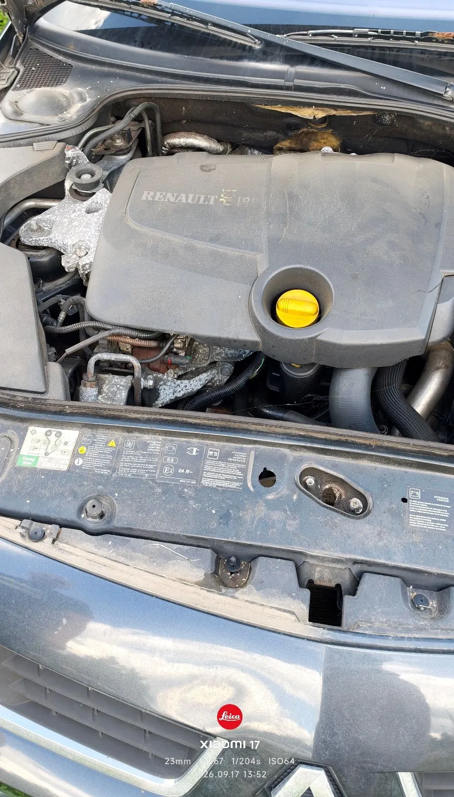 Zdjęcie przedstawiające komorę silnika samochodu marki Renault. Widoczna jest plastikowa osłona silnika z napisem 'RENAULT' oraz żółtym korkiem wlewu oleju. Poniżej znajduje się metalowy element nadwozia z naklejką techniczną zawierającą symbole i napisy, w tym '24 R-'. Na samym dole zdjęcia widoczny jest fragment przedniego zderzaka w kolorze szarym oraz znak wodny z informacjami o aparacie: 'Leica XIAOMI 11 17 23mm f/1.67 1/204s ISO64 26.09.17 13:52'.