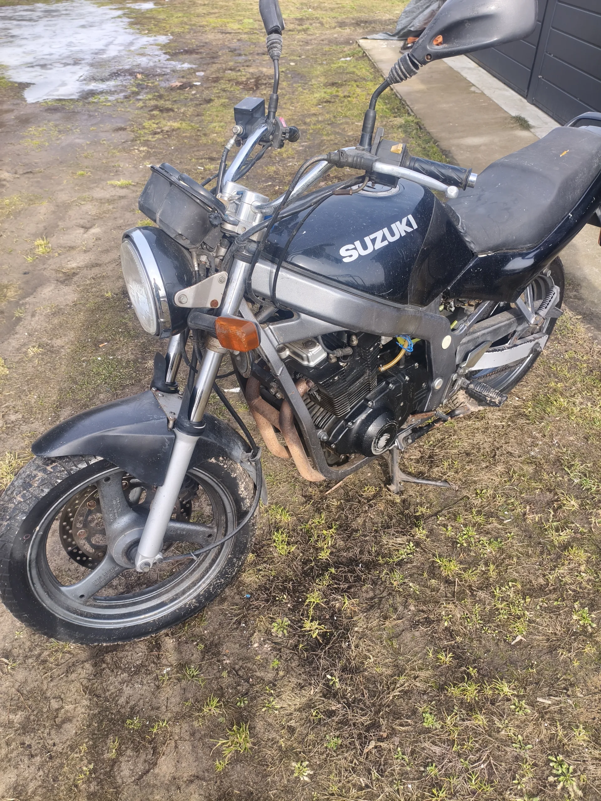 Czarny motocykl marki SUZUKI zaparkowany na trawiastym i błotnistym terenie. Widoczne są przednie koło z reflektorem, czarna siedziba, uchwyty kierownicy z lusterkami oraz silnik. W tle betonowa ścieżka, ciemny budynek i kałuża wody. Tekst SUZUKI na zbiorniku paliwa jest czytelny.