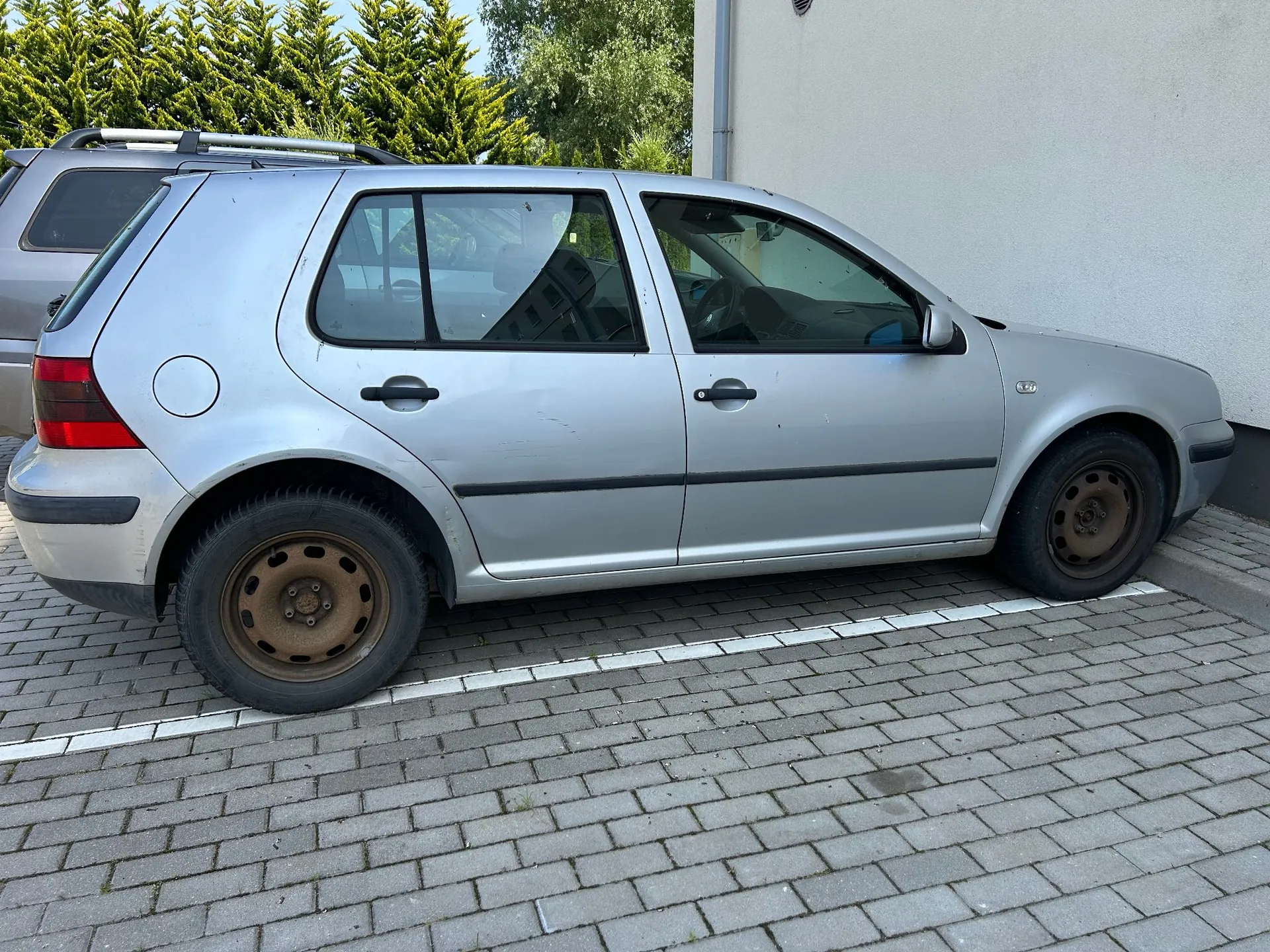 Srebrny samochód osobowy marki Volkswagen Golf IV w wersji nadwozia hatchback, zaparkowany bokiem na kostce brukowej obok białej ściany budynku. Auto posiada stalowe felgi i czarne listwy ochronne na drzwiach.