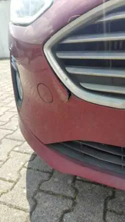 Zbliżenie na przedni zderzak czerwonego samochodu. Widoczny jest fragment grilla z poziomymi srebrnymi listwami oraz uszkodzenie w postaci pęknięcia i odkształcenia lakieru w miejscu łączenia zderzaka z ramką grilla. Samochód stoi na kostce brukowej.