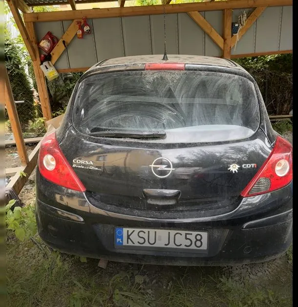 Tył czarnego, zakurzonego samochodu marki Opel Corsa z napisem CDTI. Pojazd posiada polską tablicę rejestracyjną o numerze KSU JC58. Samochód znajduje się pod drewnianą wiatą.