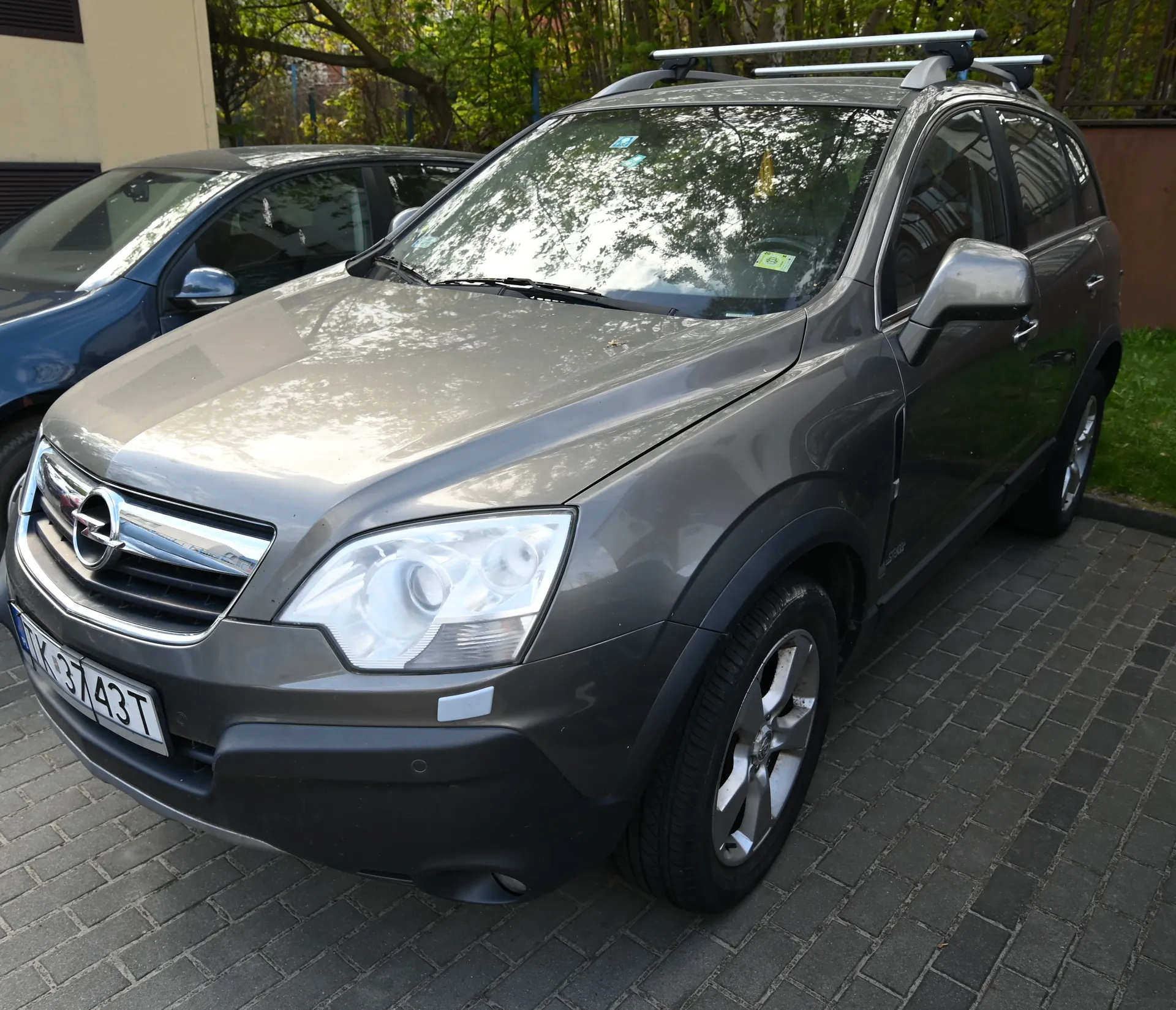 Srebrny SUV marki Opel z białą tablicą rejestracyjną VK 3743T zaparkowany na brukowanej powierzchni. Samochód posiada dachowy bagażnik i reflektory z przodu. W tle widoczny jest niebieski samochód osobowy oraz drzewa i budynek. Na przedniej szybie znajdują się naklejki.