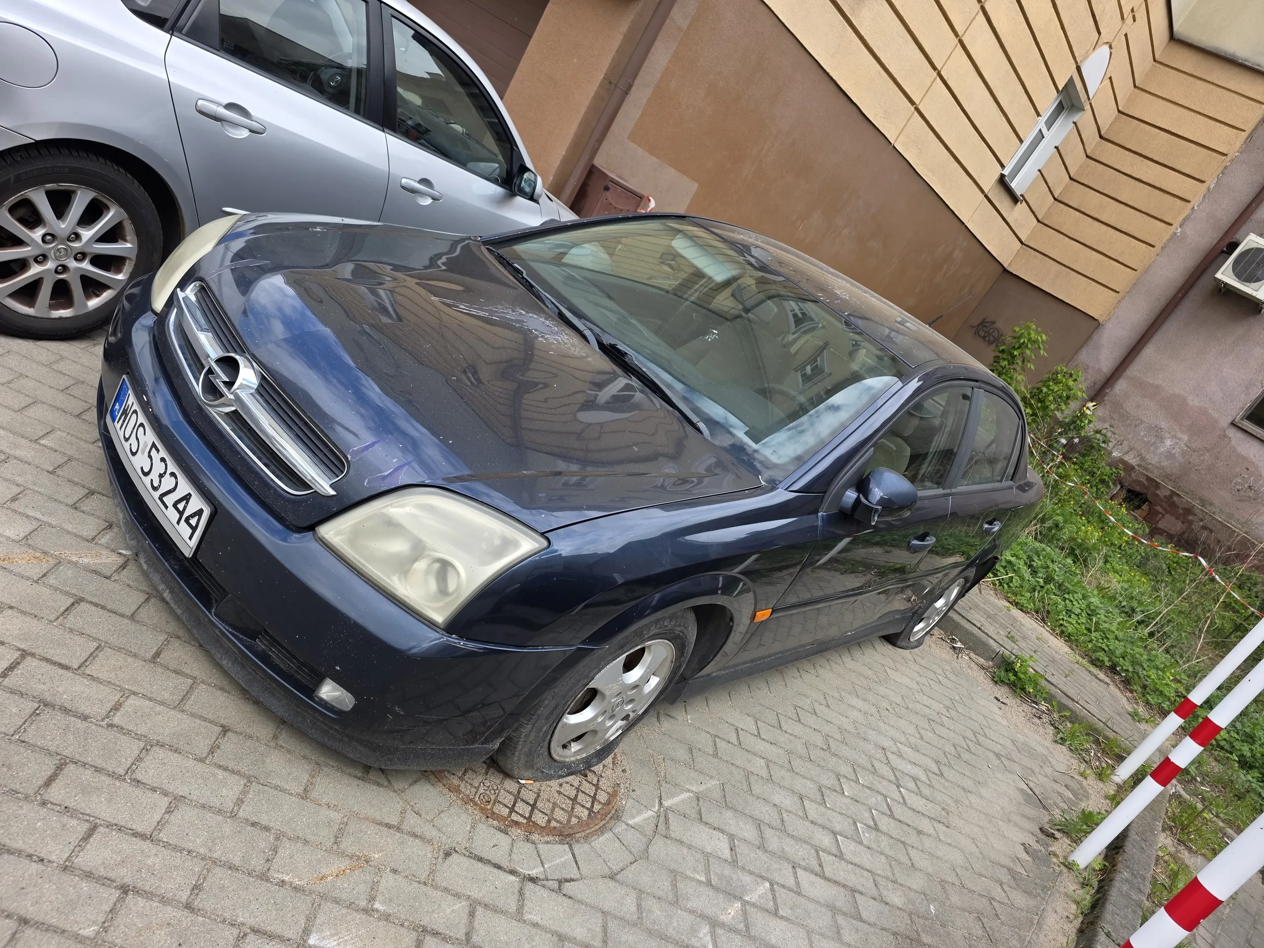 Ciemnoniebieski samochód marki Opel zaparkowany na brukowanej powierzchni. Widoczna tablica rejestracyjna z numerem WOS 53244. Obok znajduje się srebrny samochód. Po lewej stronie obrazu biało-czerwone słupy barierowe oraz zieleń. W tle fragment budynku z beżową elewacją.