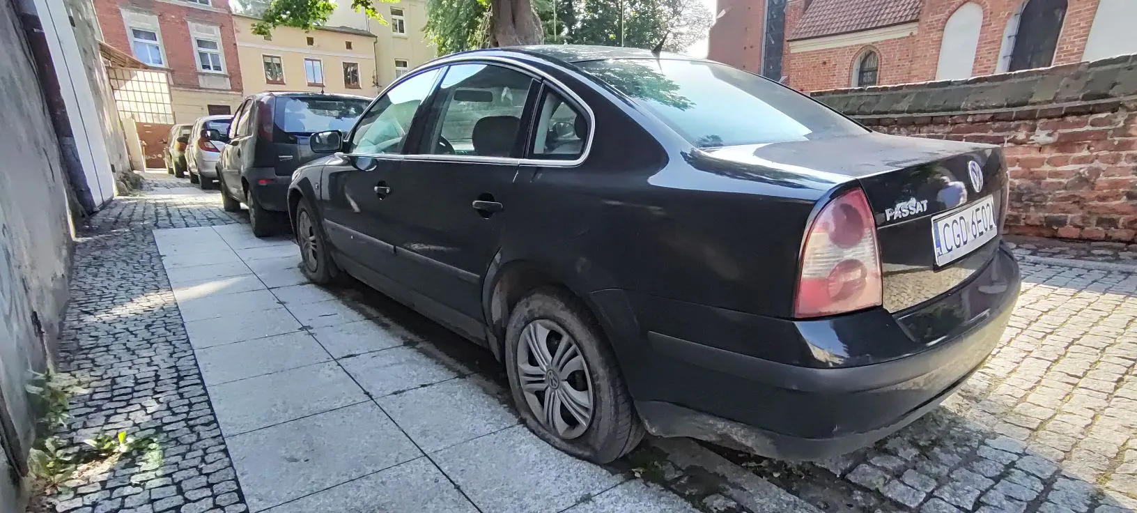 Czarny samochód osobowy marki Volkswagen Passat zaparkowany na chodniku z kostki brukowej i płyt betonowych. Auto jest widoczne z tyłu i z boku. Na tylnej klapie znajduje się napis 'PASSAT' oraz tablica rejestracyjna o numerze 'CGD 6502'. W tle widoczne są inne zaparkowane samochody oraz stare budynki z cegły i tynku.