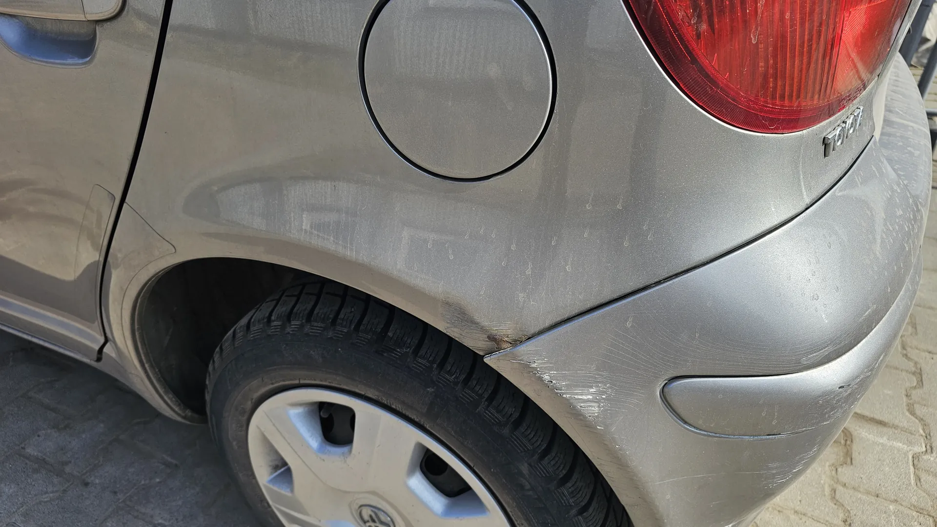 Zbliżenie na tylną część srebrnego samochodu marki Toyota. Widoczne są czerwone światło tylnego reflektora, okrągła klapa zbiornika paliwa, tylna opona z srebrną felgą oraz fragment błotnika z widocznymi zadrapaniami i zabrudzeniami. Na nadwoziu, obok reflektora, znajduje się napis 'TOYOTA'. Pod samochodem widać brukowaną nawierzchnię.