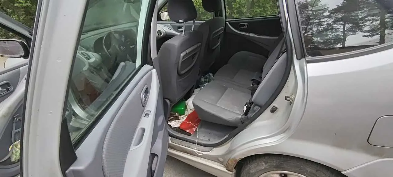 Zdjęcie przedstawia wnętrze srebrnego samochodu z otwartymi tylnymi drzwiami. Widoczne są szare siedzenia, na podłodze leżą różne przedmioty: zielona butelka, czerwony kawałek materiału oraz inne śmieci. W tle przez szyby widać drzewa. Samochód znajduje się na zewnątrz, a jego karoseria wydaje się nieco zabrudzona.