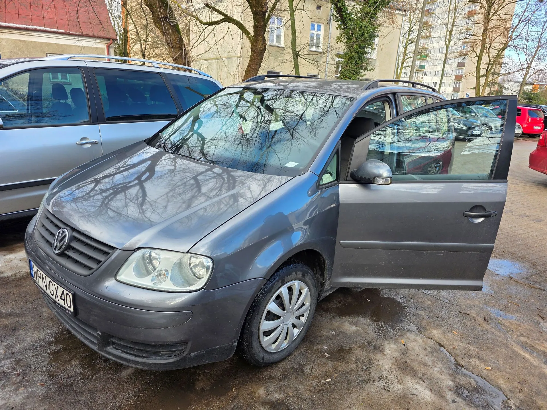 Srebrzysto-szary samochód marki Volkswagen Touran zaparkowany na błotnistym placu. Przednie prawe drzwi pasażera są otwarte. Samochód ma tablicę rejestracyjną o numerze WPN GX 40. W tle widoczny jest inny srebrny samochód, drzewa bez liści oraz jasny budynek mieszkalny.