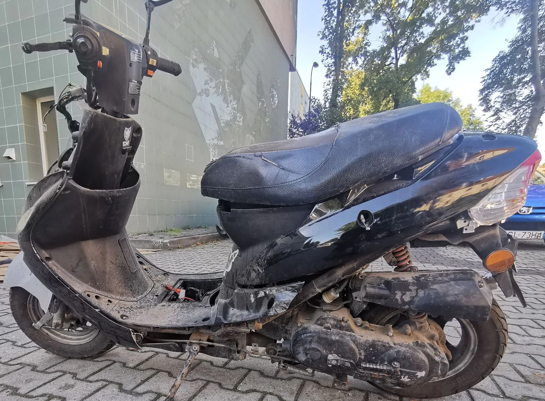 Czarny skuter zaparkowany na kostce brukowej przed szarym budynkiem. Pojazd jest w złym stanie technicznym: brakuje przedniej owiewki, siedzenie jest przetarte i zniszczone, a dolne partie silnika i ramy są zabrudzone. W tle widoczne są drzewa i fragment niebieskiego samochodu z tablicą rejestracyjną zaczynającą się od liter GL.