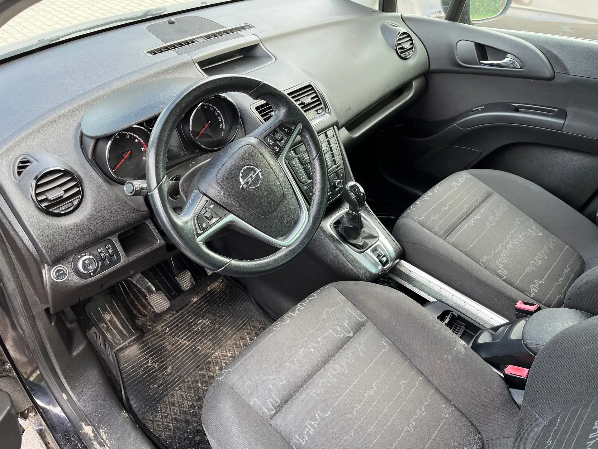 Wnętrze samochodu marki Opel z perspektywy kierowcy. Widoczna jest czarna kierownica z logo producenta, deska rozdzielcza z analogowymi wskaźnikami prędkości i obrotów, lewarek zmiany biegów oraz dwa przednie fotele z ciemnoszarą tapicerką w delikatne wzory. Na podłodze znajdują się gumowe dywaniki.