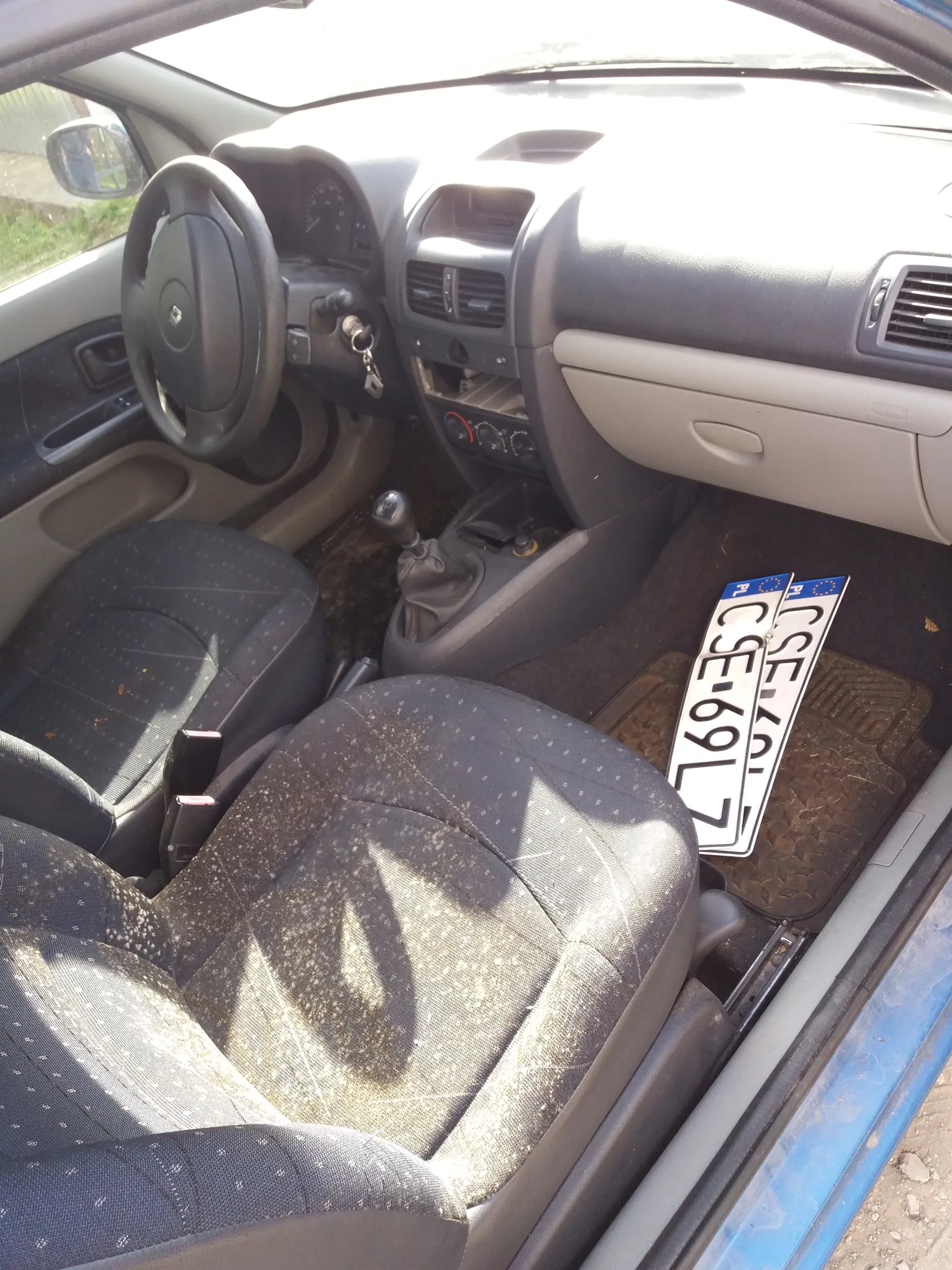 Wnętrze samochodu z widocznym kierownicą po lewej stronie, deska rozdzielcza z wentylacjami, dźwignią zmiany biegów oraz dwoma polskimi tablicami rejestracyjnymi CSE-69L7 leżącymi na dywaniku podłogowym. Siedzenia pokryte tkaniną z drobnymi plamami, fragment niebieskiego nadwozia widoczny przez okno. Na tablicach widoczne oznaczenie PL oraz czarny tekst CSE-69L7.