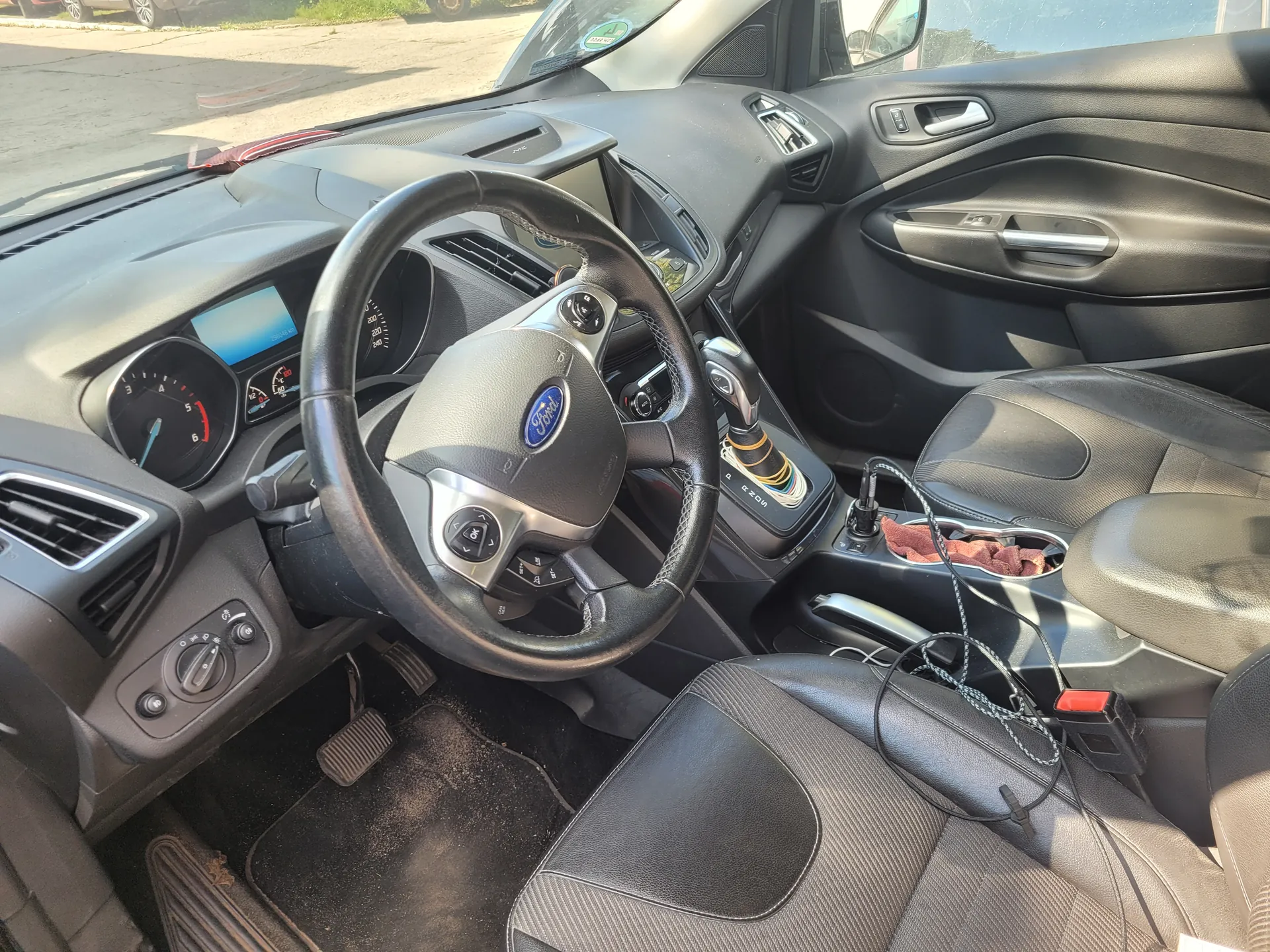 Wnętrze samochodu marki Ford z widocznym kierownicą z logo Ford, tablicą rozdzielczą z analogowymi wskaźnikami i cyfrowym wyświetlaczem, centralną konsolą z dźwignią zmiany biegów, czarnymi skórzonymi fotelami oraz przedmiotami takimi jak czerwona szmatka w uchwycie na napoje i przewody elektryczne. Przez przednią szybę widoczna jest ulica z innymi pojazdami i zielenią.