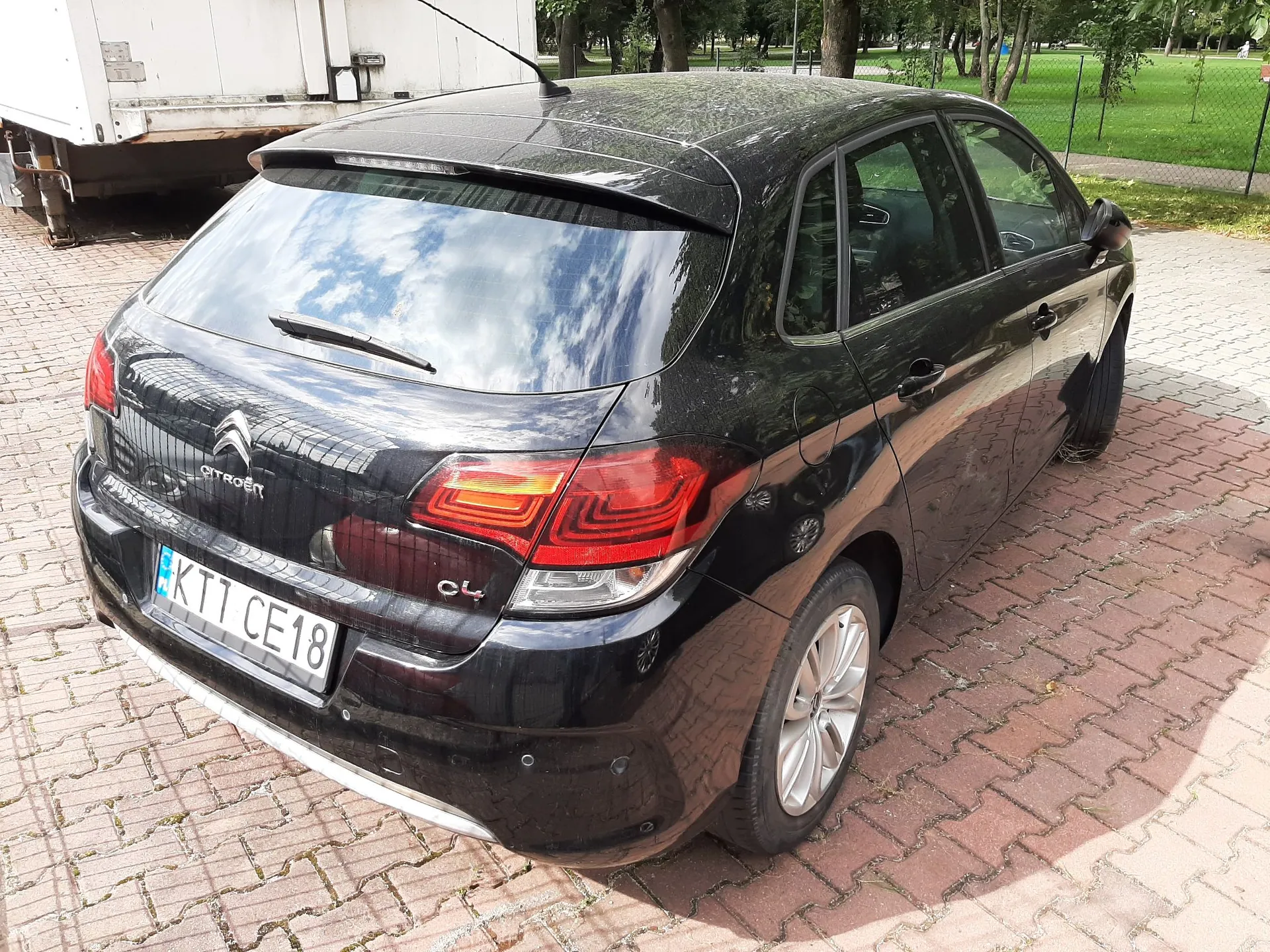 Czarny samochód marki Citroën model C4 zaparkowany na kostce brukowej. Widok z tyłu i z boku. Samochód ma polską tablicę rejestracyjną o numerze KTI CE18. W tle widoczny jest biały pojazd ciężarowy oraz zielony obszar z drzewami.