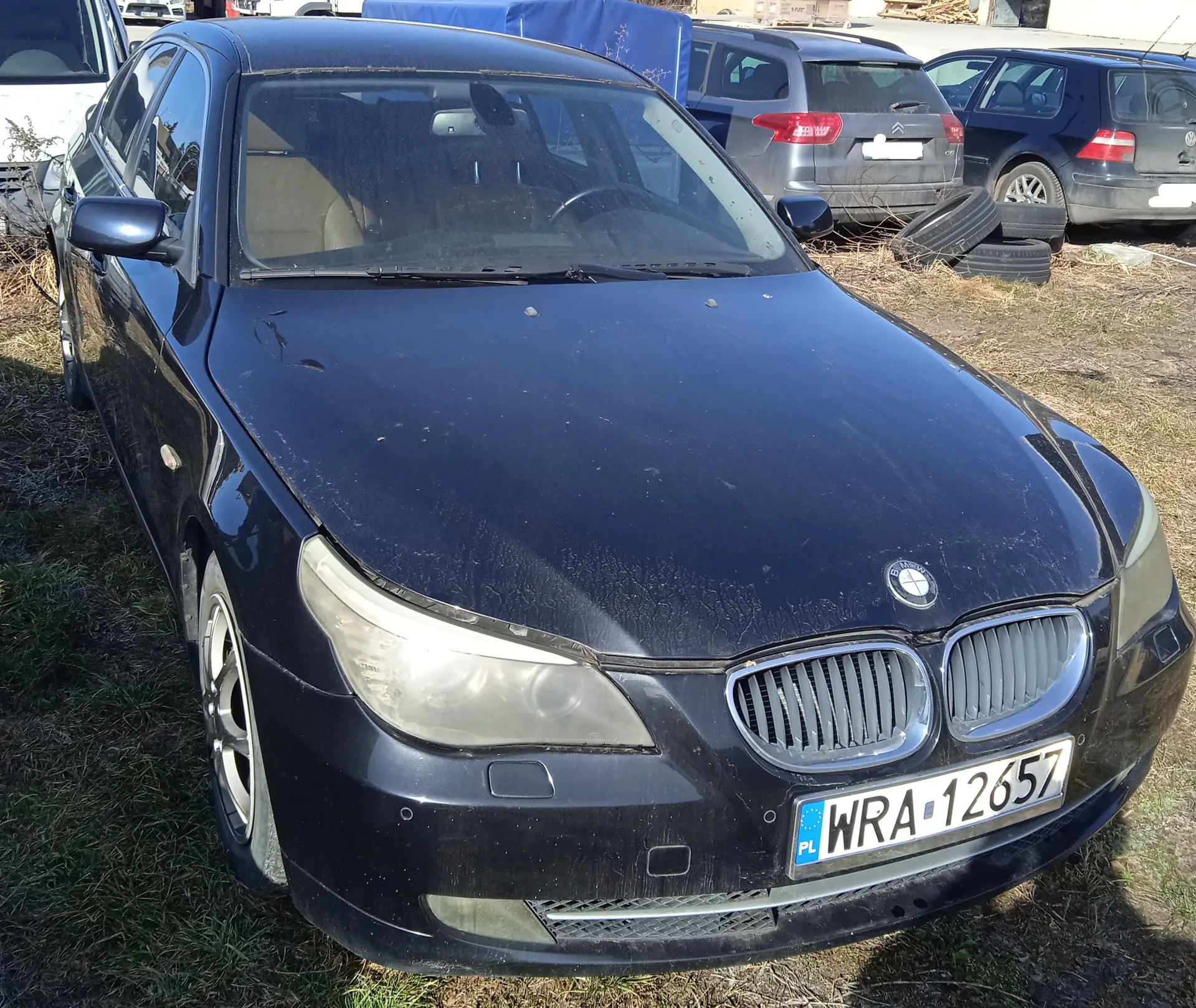 Czarny samochód marki BMW z przodu, z widoczną tablicą rejestracyjną PL WRA-12657. Samochód stoi na trawie, w tle znajdują się inne pojazdy: szary Citroën i ciemny Volkswagen, a także stos opon. Na masce BMW widoczny jest logo marki, a samochód ma ślady zabrudzeń i zadrapań. Tło obejmuje suchą trawę i fragmenty innych pojazdów.