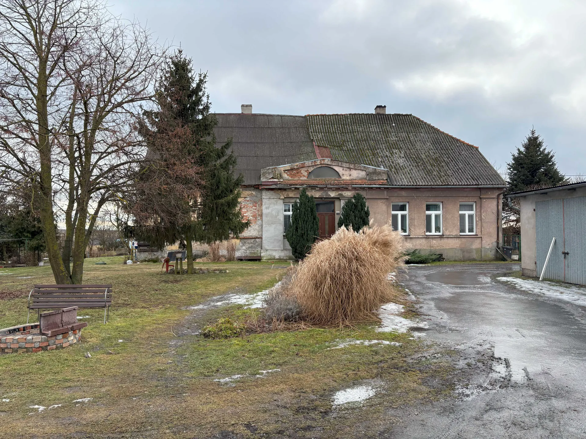 Stary dom jednorodzinny z uszkodzonym dachem krytym blachą, częściowo zarośniętym mchem. Dom ma brązowe drzwi, białe okna i fragmenty ceglanej elewacji. Przed domem znajduje się duża, sucha kępka trawy, ławka, ceglany obiekt oraz drzewa bez liści i iglaste. Po prawej stronie widoczny garaż z metalowymi drzwiami. Na trawniku i nawierzchni drogi pozostałości śniegu, niebo zachmurzone. W tle drzewa i niewielkie budowle.