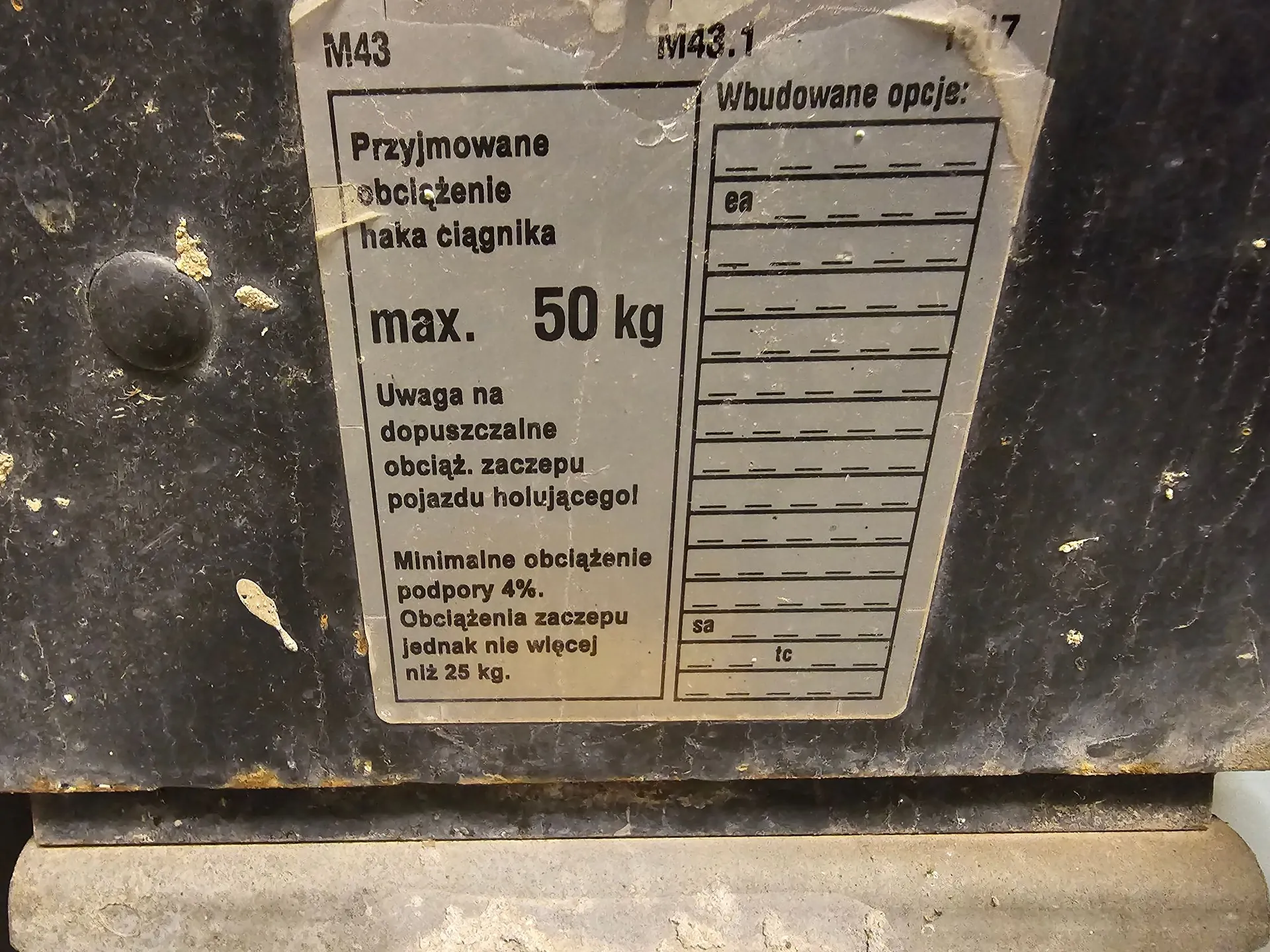 Zdjęcie tabliczki znamionowej przymocowanej do ciemnej, metalowej powierzchni. Tabliczka zawiera tekst w języku polskim: 'M43 M43.1', 'Przyjmowane obciążenie haka ciągnika max. 50 kg', 'Uwaga na dopuszczalne obciąż. zaczepu pojazdu holującego!', 'Minimalne obciążenie podpory 4%. Obciążenia zaczepu jednak nie więcej niż 25 kg.'. Po prawej stronie znajduje się tabela z nagłówkiem 'Wbudowane opcje:' z pustymi polami i oznaczeniami 'ea', 'sa', 'ic'.