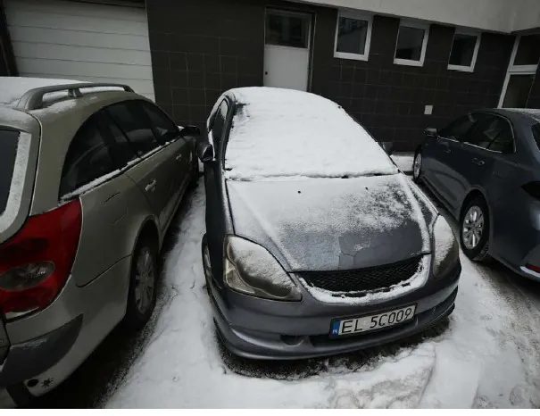Trzy samochody zaparkowane na śnieżnej powierzchni. W centrum szary sedan z pokrytą śniegiem maską i tablicą rejestracyjną EL 5C009. Po lewej srebrny kombi z dachowym bagażnikiem, po prawej ciemnoszary sedan. Tło stanowi budynek z ciemnoszarymi płytkami, białymi oknami i bramą garażową. Śnieg pokrywa samochody i ziemię.