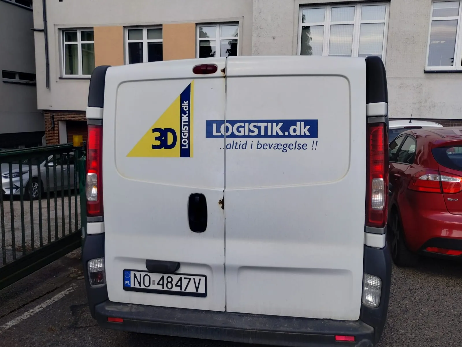 Tył białego furgonu z logo w kształcie żółtego trójkąta z napisem 3D oraz pionową niebieską listwą z tekstem LOGISTIK.dk. Po prawej stronie drzwi furgonu niebieski prostokąt z napisem LOGISTIK.dk i pod nim tekstem ...altid i bevægelse !!. Tablica rejestracyjna z polskim kodem PL i numerem NO 4847V. W tle budynek z oknami, czerwony samochód osobowy oraz zielone ogrodzenie. Inne pojazdy widoczne po lewej stronie.