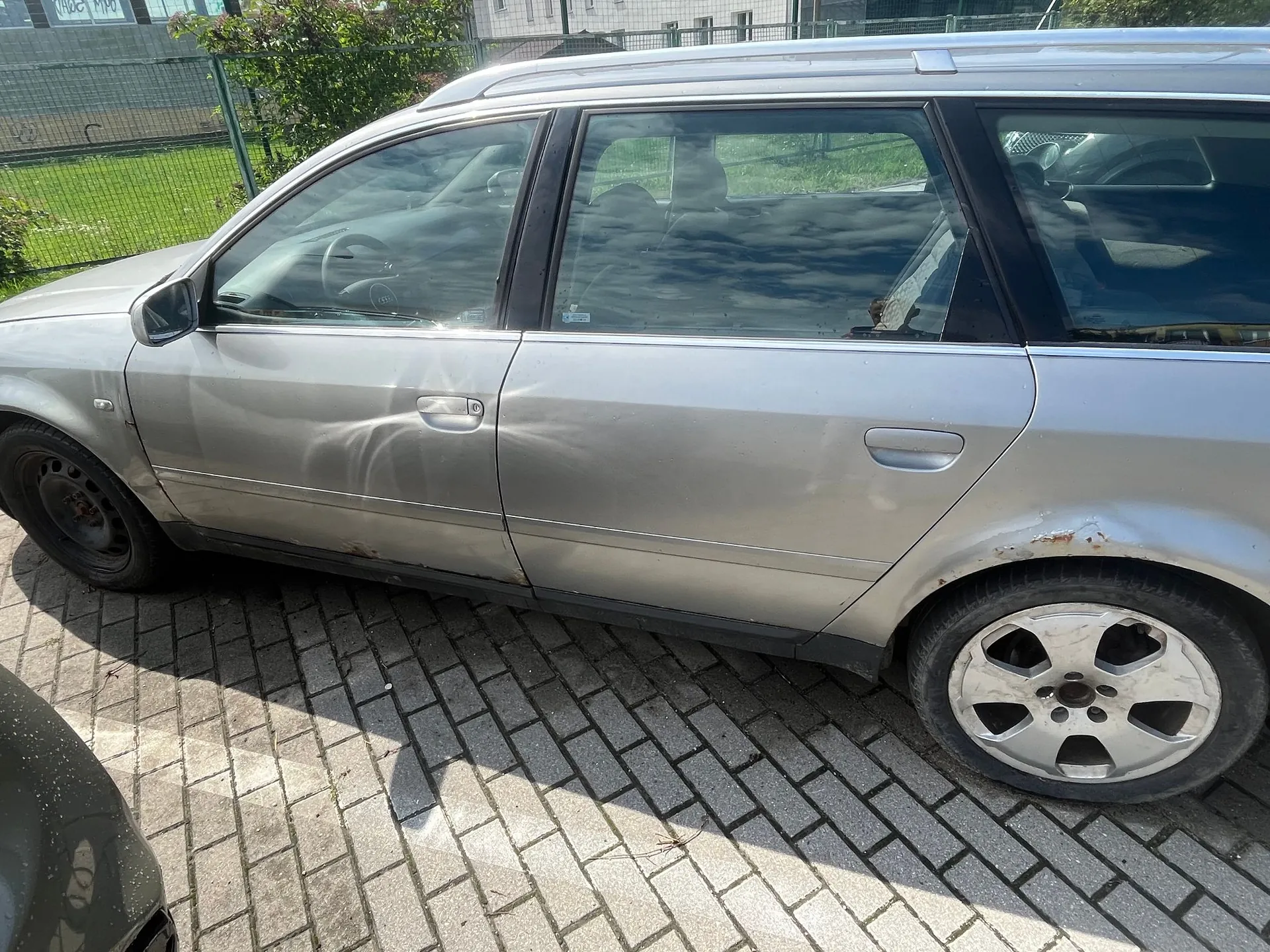 Srebrny samochód typu kombi zaparkowany na kostce brukowej. Pojazd posiada widoczne uszkodzenia karoserii w postaci wgnieceń na przednich i tylnych drzwiach od strony pasażera oraz ślady korozji na dolnej krawędzi nadwozia i nad tylnym kołem.