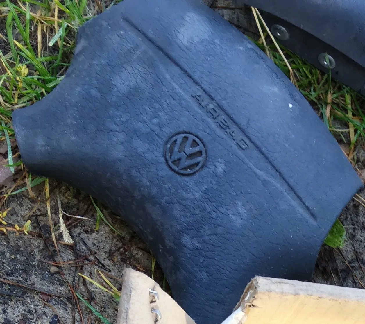 Zbliżenie na ciemnoszarą, gumową lub plastikową osłonę samochodową leżącą na ziemi z trawą. Na powierzchni elementu znajduje się wytłoczone logo marki Volkswagen oraz napis 'AIRBAG'.