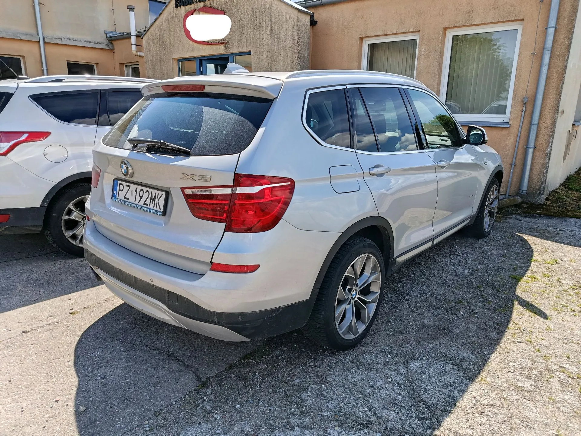 Srebrny samochód marki BMW X3 zaparkowany na betonowym podjeździe przed budynkami. Widoczna jest tylna część i prawy bok pojazdu. Samochód posiada polską tablicę rejestracyjną o numerze PZ 192MK. W tle po lewej stronie widoczny jest fragment innego, białego samochodu.