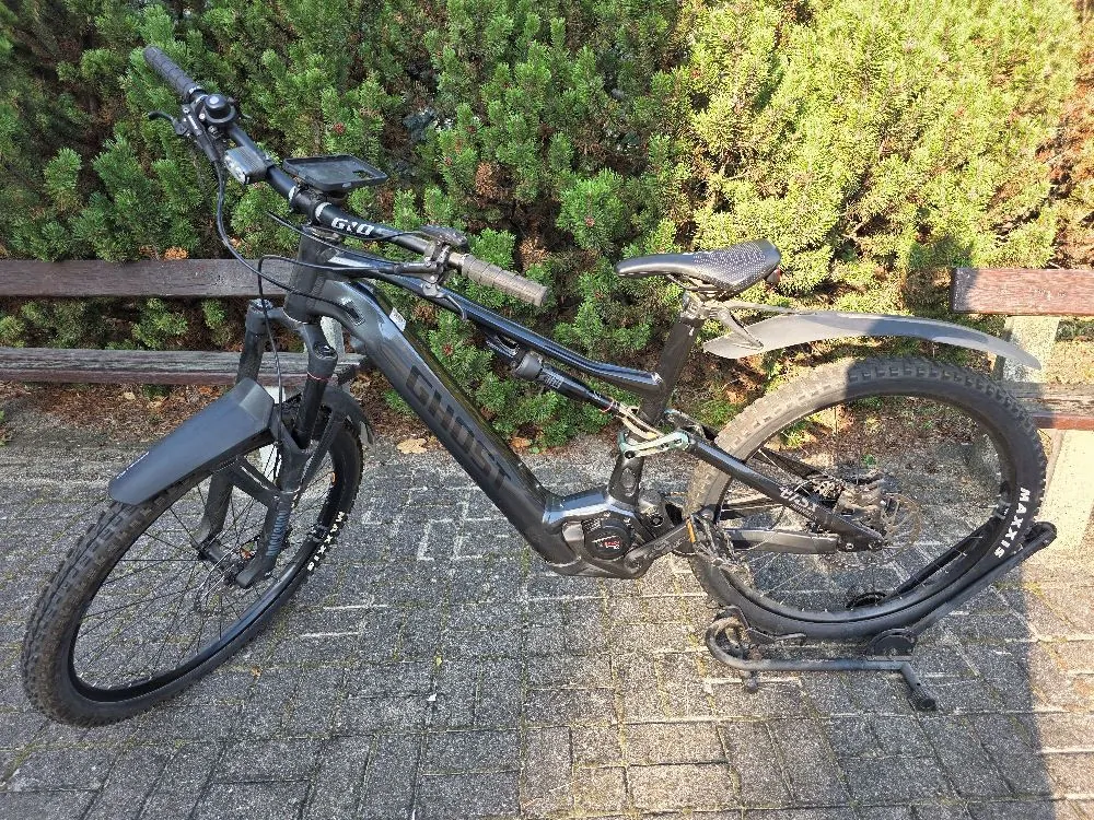 Czarny rower elektryczny marki Ghost, ustawiony bokiem na kostce brukowej. Rower posiada amortyzator przedni i tylny, szerokie opony Maxxis oraz błotniki. W tle widoczna jest zielona roślinność i drewniana ławka.