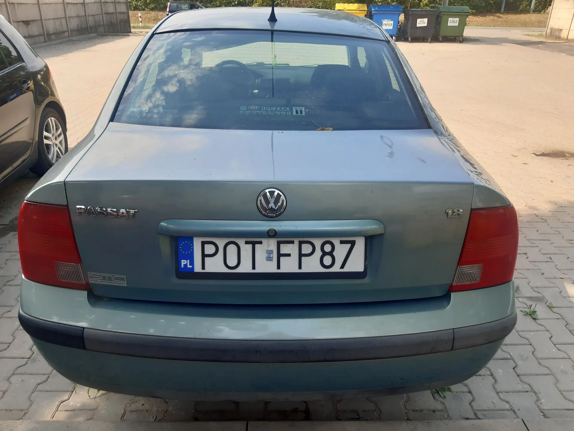 Tylny widok jasnoniebieskiego samochodu marki Volkswagen Passat. Na klapie bagażnika znajduje się logo Volkswagena, napis 'PASSAT' po lewej stronie oraz oznaczenie '1.8' po prawej. Samochód posiada polską tablicę rejestracyjną o numerze POT FP87. Auto zaparkowane jest na kostce brukowej.