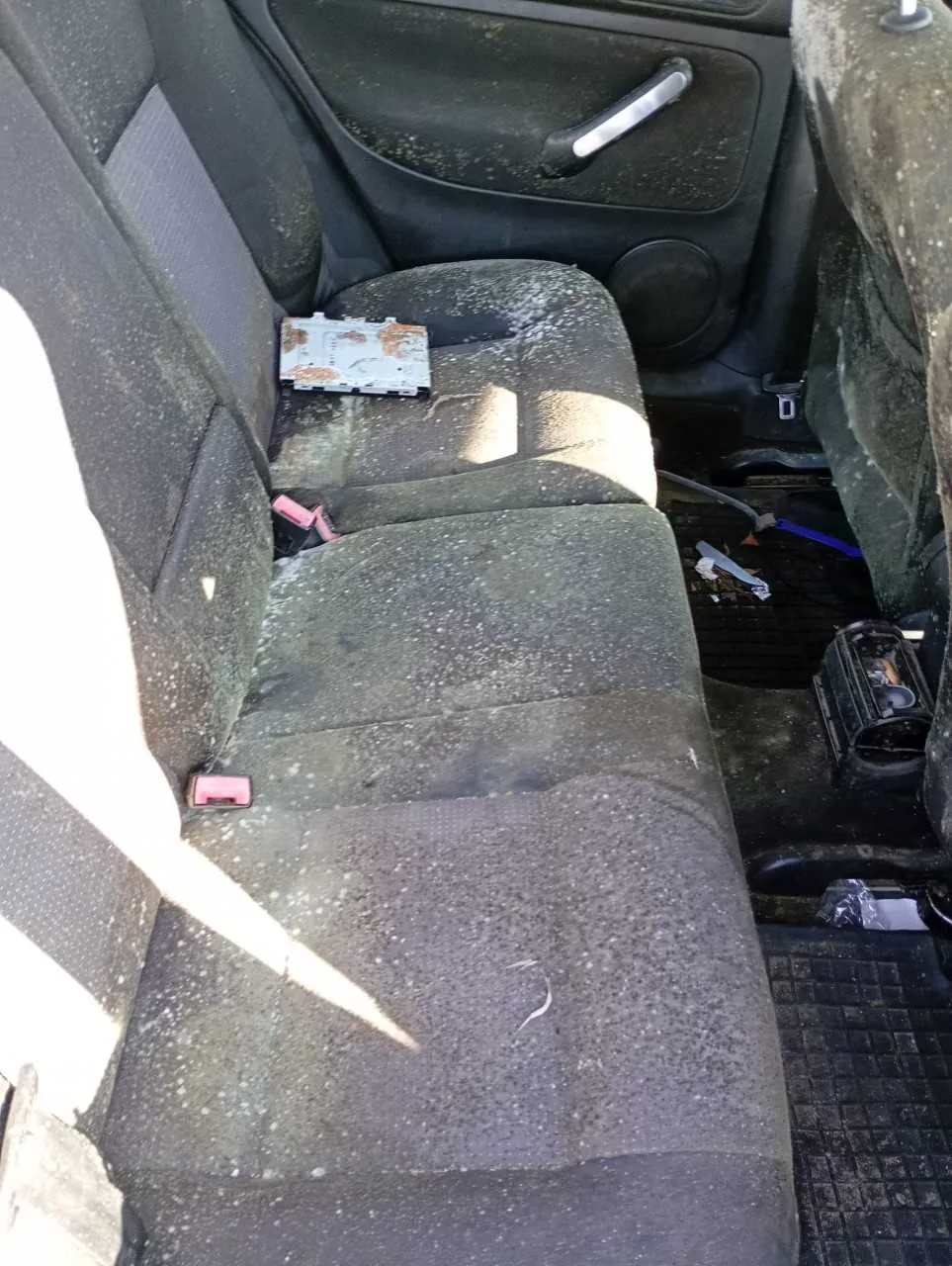 Wnętrze samochodu z widocznym tylnym siedzeniem pokrytym białymi plamami i brudem. Na siedzeniu leży zardzewiały lub uszkodzony prostokątny przedmiot. Widoczne są czarne poduszki siedzeń z czerwonymi elementami pasów bezpieczeństwa. Drzwi pasażerskie z czarną obudową, uchwytem i głośnikiem. Na podłodze znajdują się różne przedmioty, w tym niebieski element i czarny pojemnik. Całość w stanie nieporządku i zanieczyszczenia.