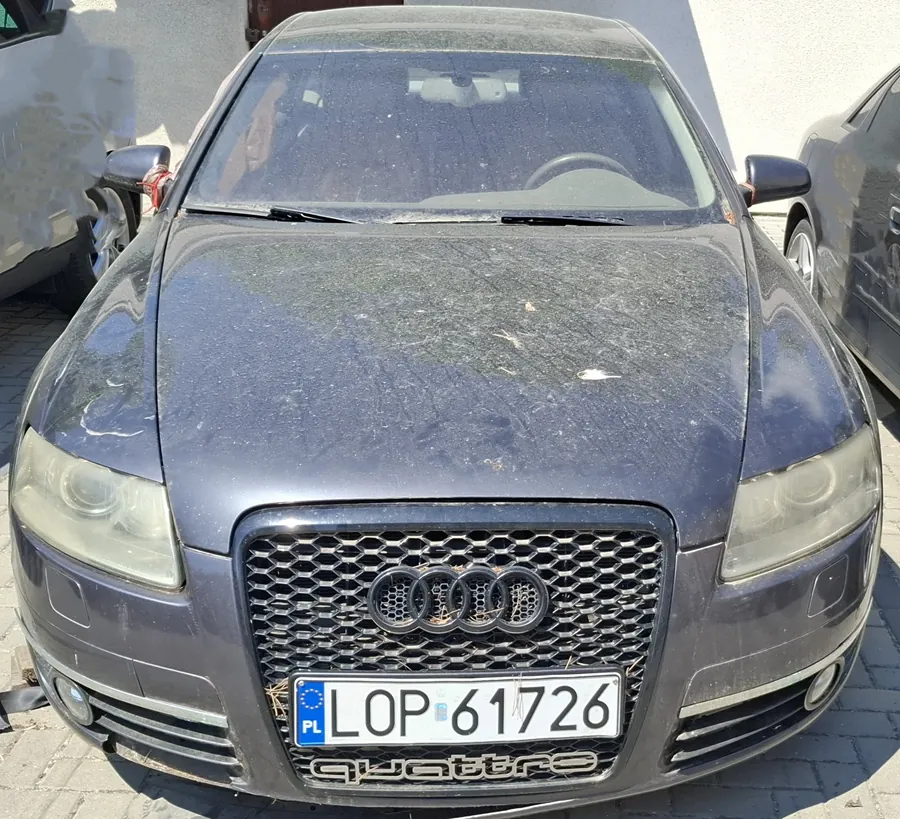 Czarny samochód Audi z przodu, z widocznym logo Audi na atrapie chłodnicy i napisem 'quattro' poniżej. Na masce brud oraz resztki liści. Tablica rejestracyjna PL LOP 61726. Po obu stronach częściowo widoczne inne samochody. Tło: biała ściana budynku i brukowana powierzchnia.