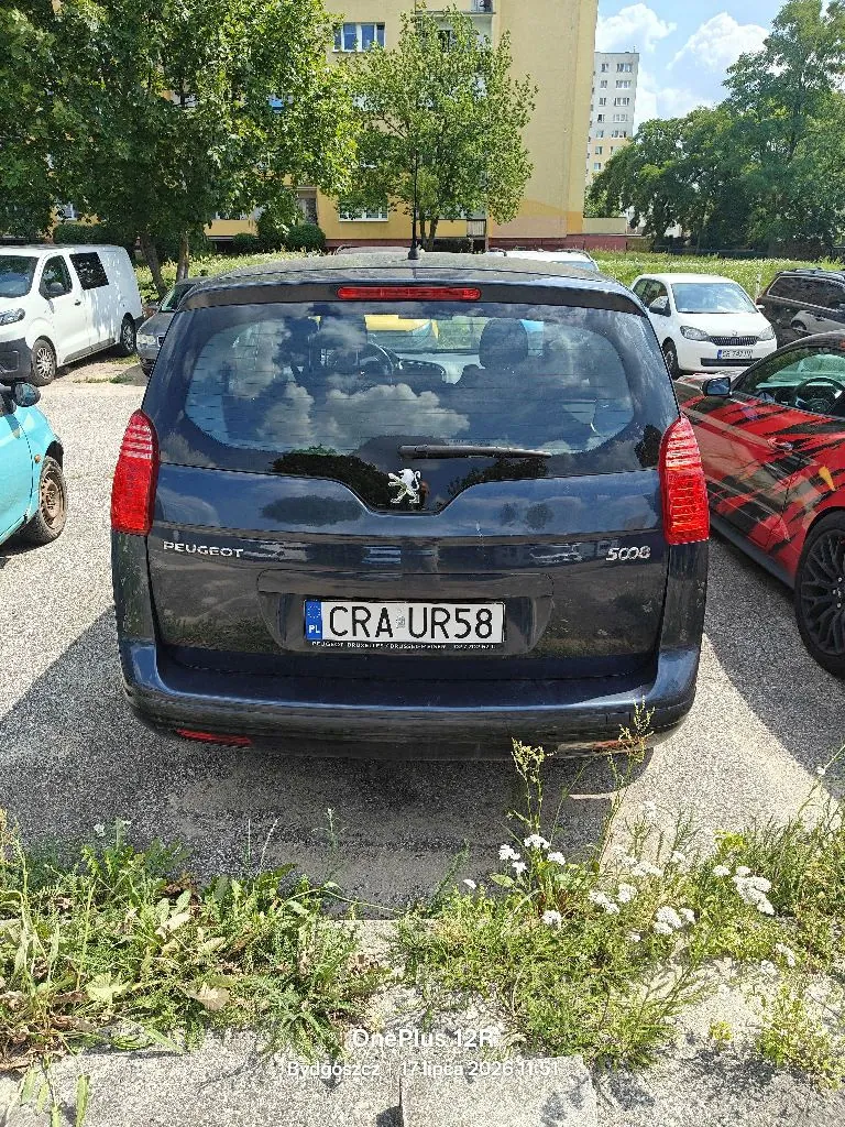 Tył ciemnoniebieskiego samochodu marki Peugeot 5008 zaparkowanego na parkingu. Pojazd ma polską tablicę rejestracyjną o numerze CRA UR58. W tle widoczne są inne samochody, drzewa oraz fragment żółtego budynku mieszkalnego. Na dole zdjęcia znajduje się znak wodny z tekstem 'OnePlus 12R Bydgoszcz 17 lipca 2024 11:51'.