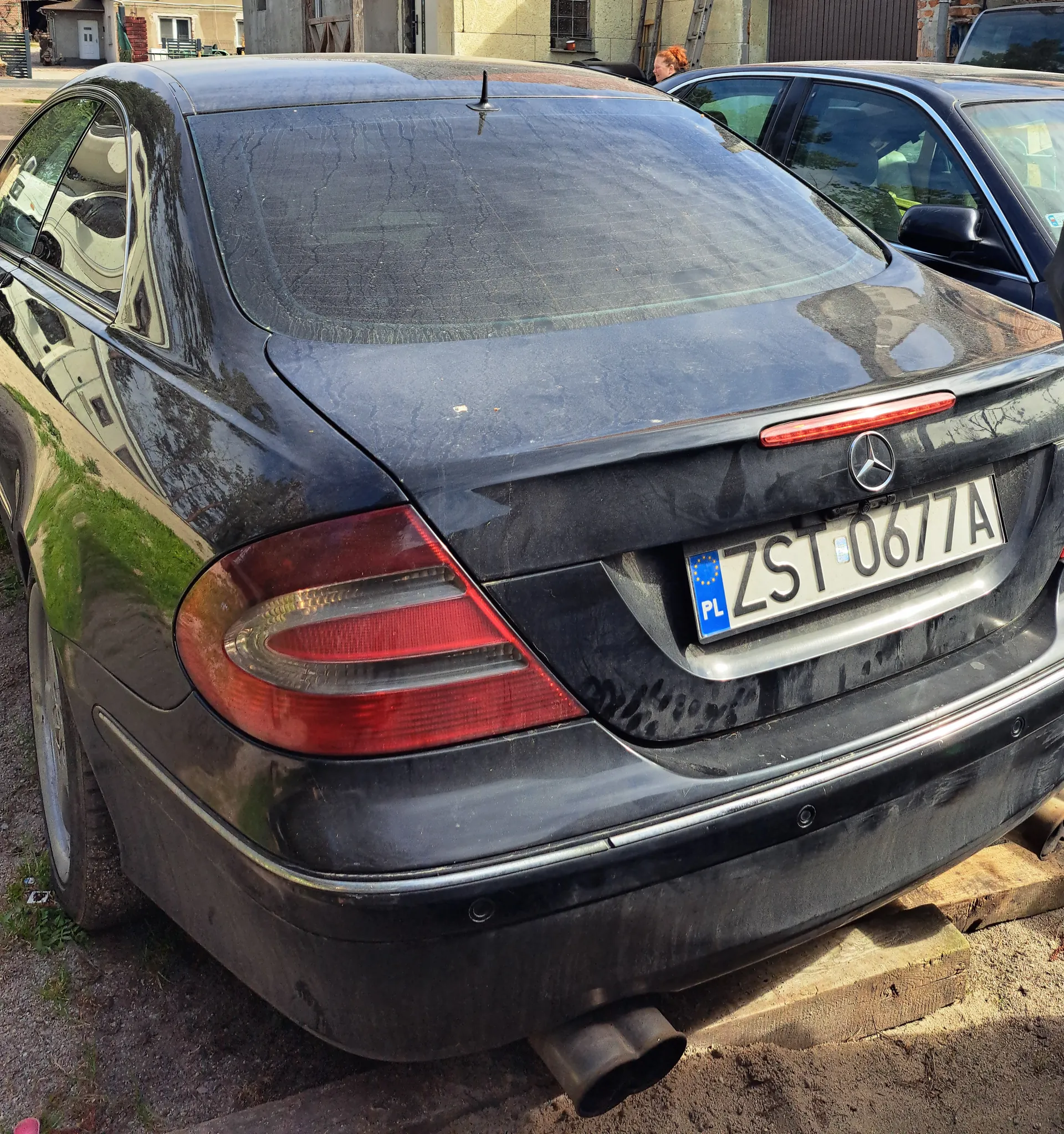 Tył czarnego samochodu marki Mercedes-Benz z tablicą rejestracyjną ZST 0677A. Tablica posiada niebieski pas z napisem PL i symbolem Unii Europejskiej. Samochód jest zabrudzony, widoczne są czerwone światła tylnego oraz wydech. W tle drugi czarny samochód, budynki, drabina oraz osoba z rudymi włosami. Podwozie samochodu opiera się na drewnianych blokach na podłożu z żwiru i trawy.