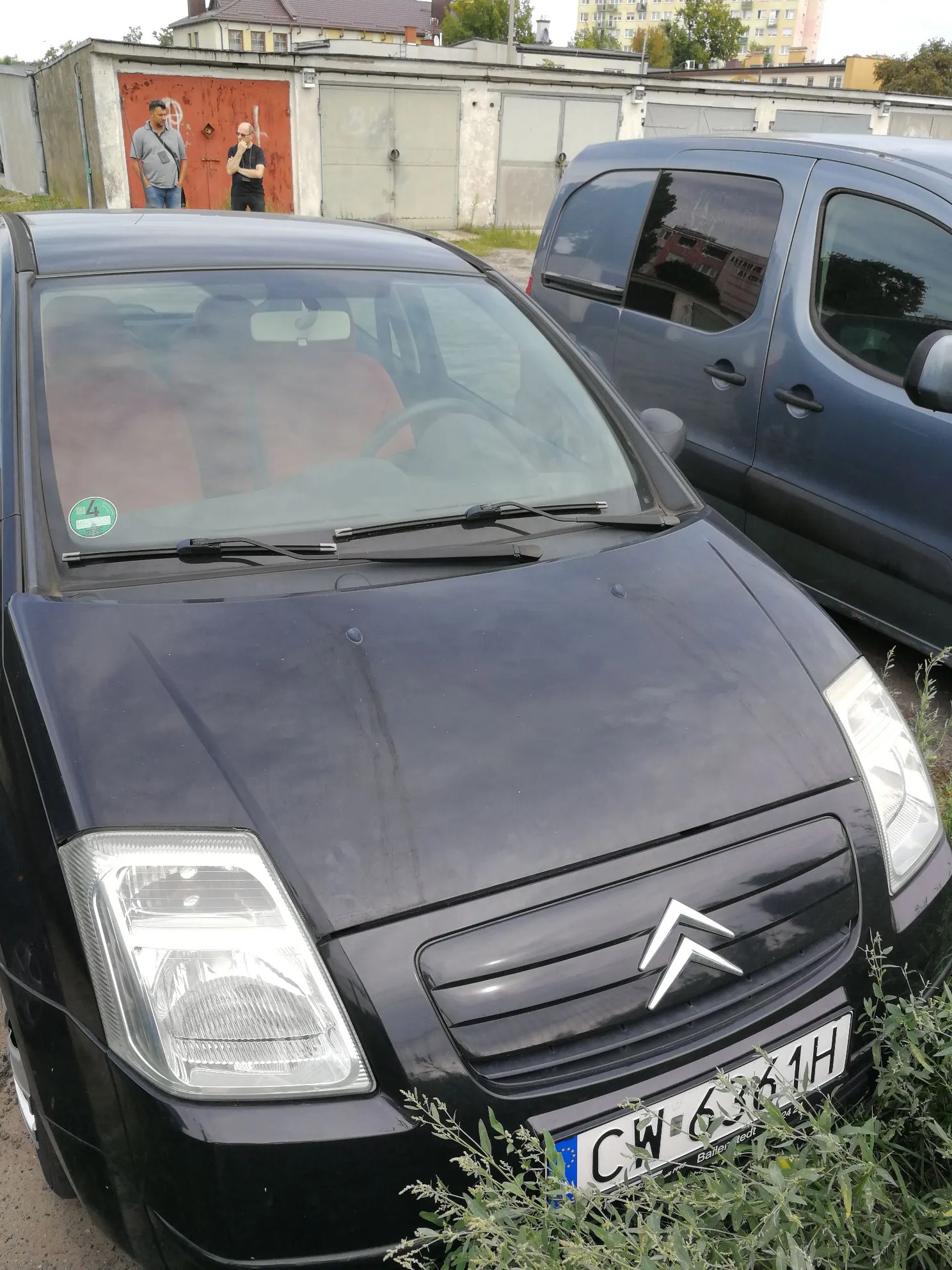 Czarny samochód marki Citroën zaparkowany obok ciemnoniebieskiego vana. Na pierwszym planie widoczny jest przód Citroëna z logo marki na grillu oraz tablicą rejestracyjną o numerze CW 6361H. W tle widać szare garaże oraz dwie osoby stojące przed nimi.