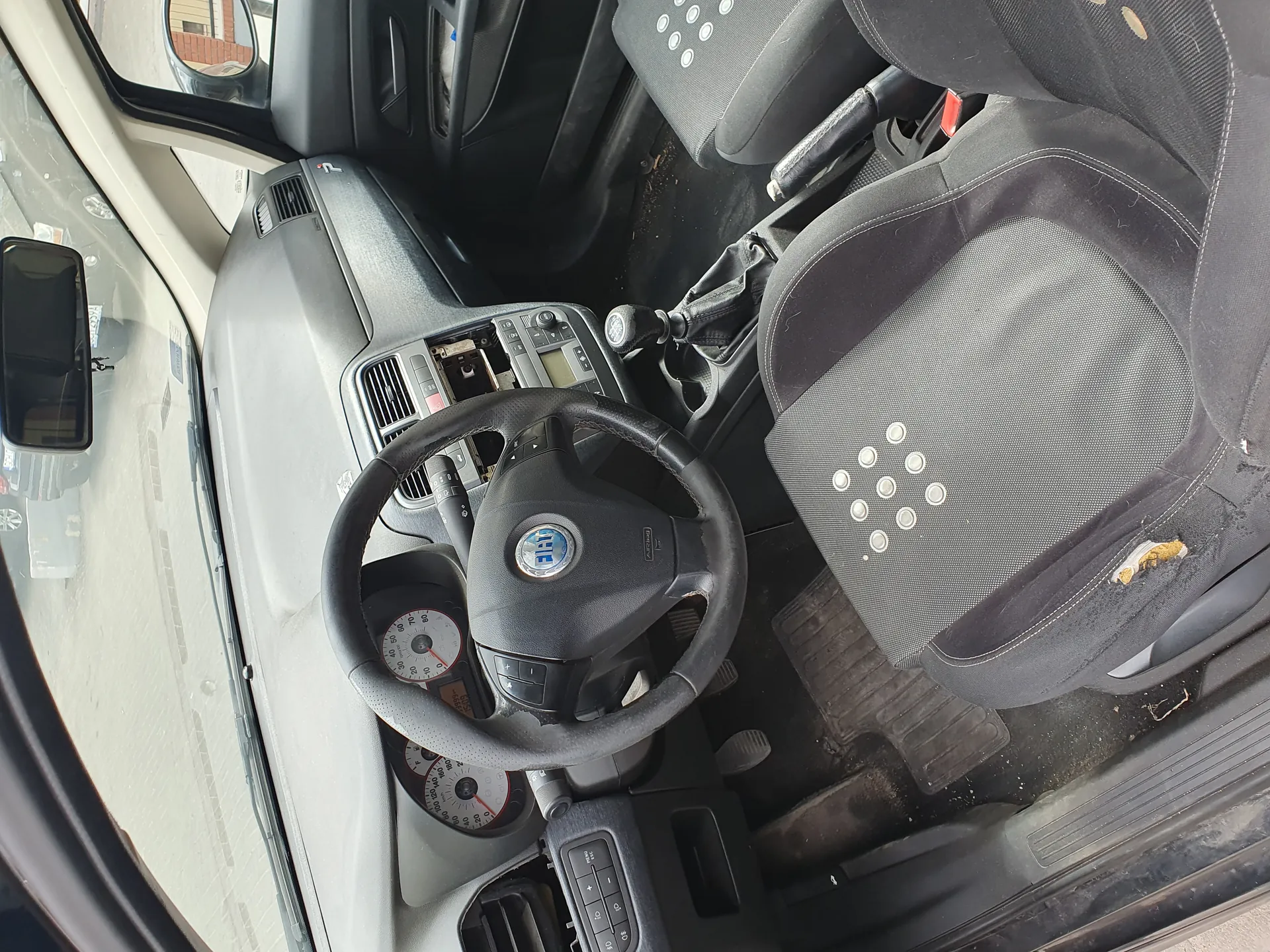 Wnętrze samochodu z kierownicą marki Fiat, tablicą rozdzielczą z licznikami prędkości i obrotomierzem, czarnymi siedzeniami z widocznymi uszkodzeniami, dźwignią zmiany biegów, pedałami oraz widokiem przez przednią szybę na budynki i inny samochód.
