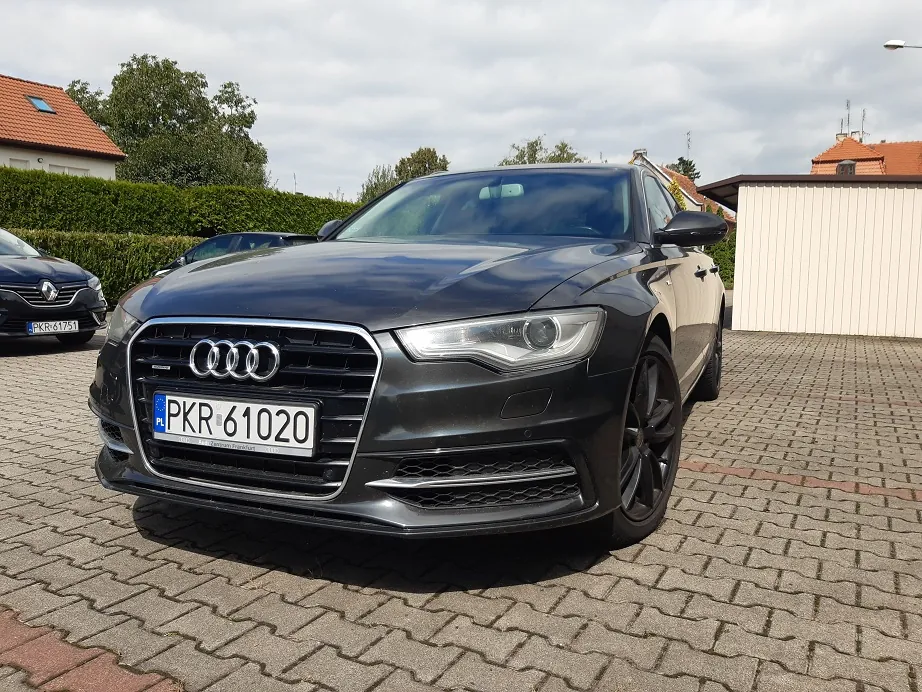 Czarny samochód marki Audi zaparkowany na kostce brukowej. Widoczny jest przód pojazdu z charakterystycznym grillem, logo Audi oraz polską tablicą rejestracyjną o numerze PKR 61020. W tle widać inny ciemny samochód, zielony żywopłot oraz fragmenty budynków z dachami krytymi czerwoną dachówką pod zachmurzonym niebem.